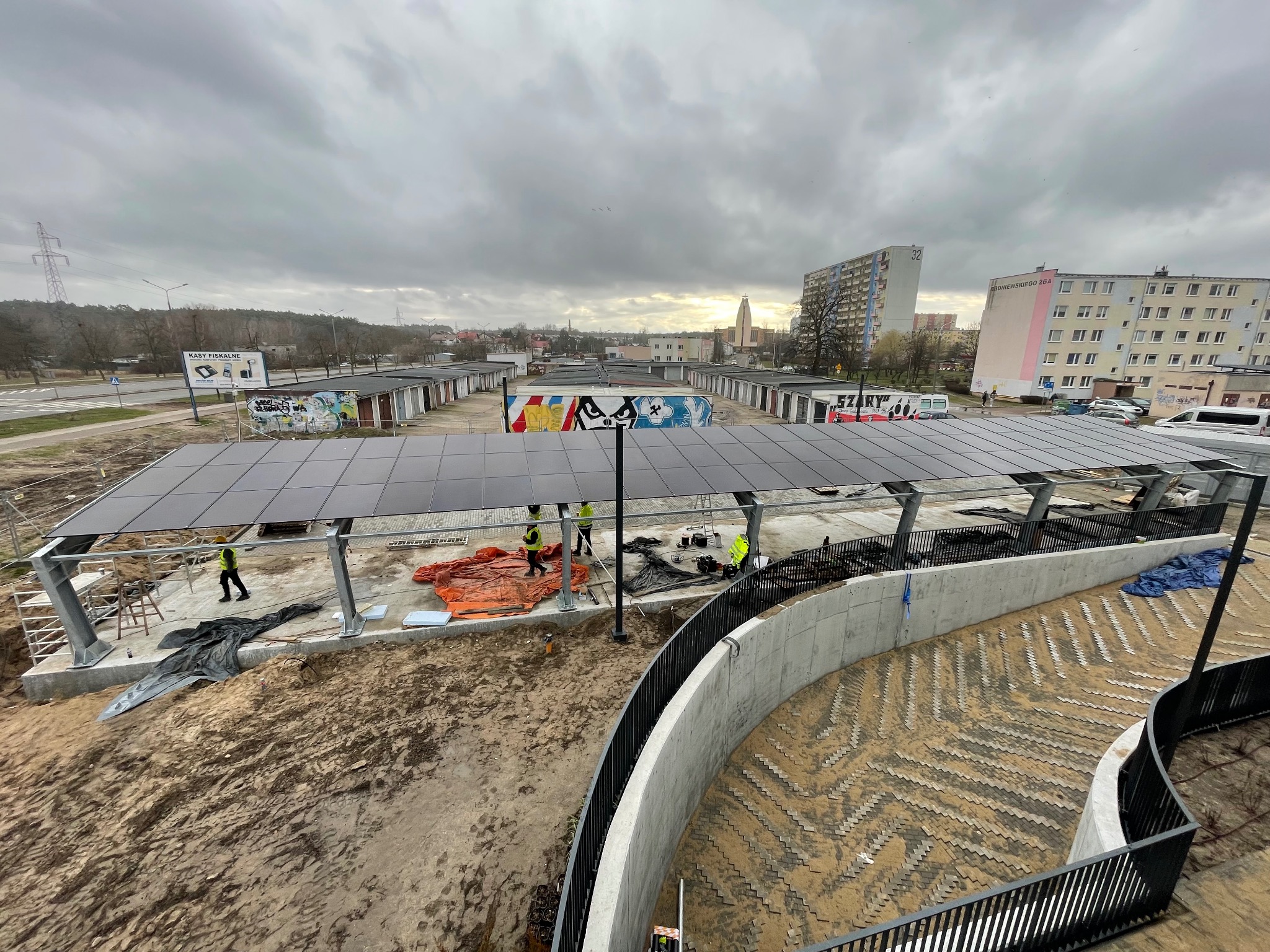 Montaż zadaszenia z paneli słonecznych nad parkingiem, widoczne elementy konstrukcyjne, pracownicy w kamizelkach odblaskowych, w tle zabudowa miejska i pochmurne niebo.