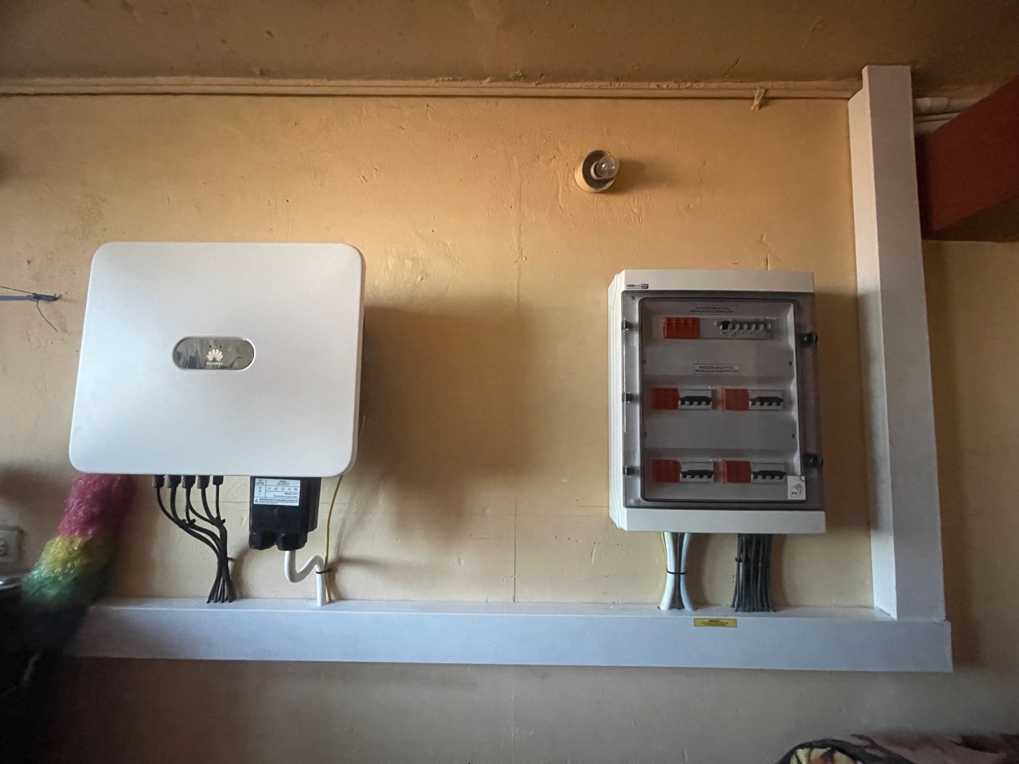 Biały inwerter Huawei i skrzynka elektryczna z zabezpieczeniami zamontowane na ścianie nad białą półką, widoczne przewody elektryczne i fragment kolorowej miotełki do kurzu.
