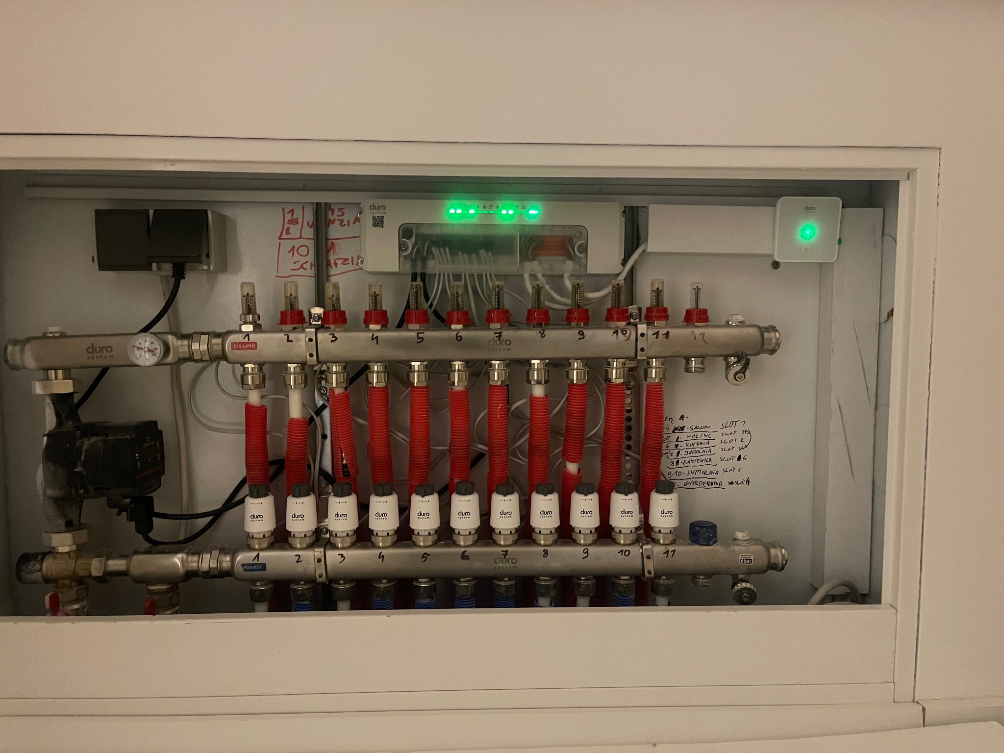 Rozdzielacz ogrzewania podłogowego w białej szafce z widocznymi rurami zasilającymi i powrotnymi, pompą obiegową, sterownikiem z diodami LED i notatkami na ścianie szafki.