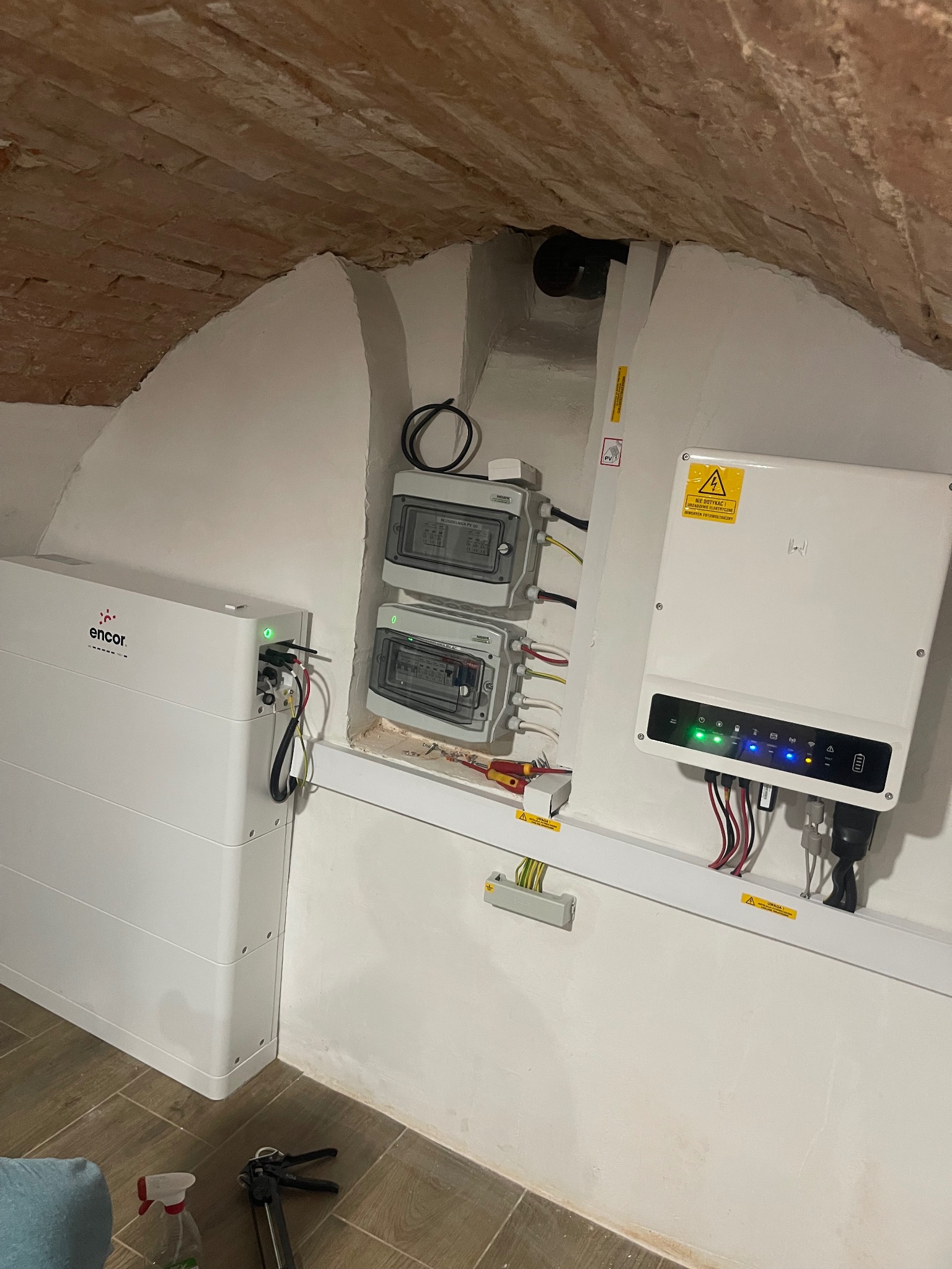 Instalacja magazynu energii Encor i inwertera w pomieszczeniu z ceglanym sklepieniem łukowym, widoczne zabezpieczenia elektryczne i narzędzia montażowe.
