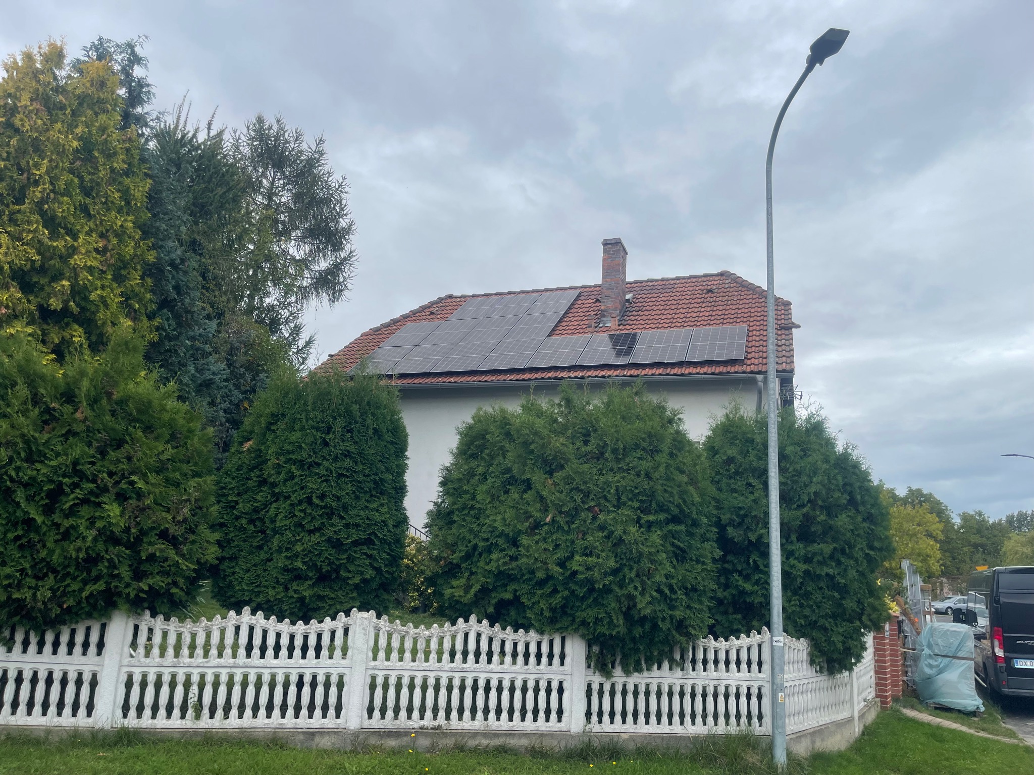 Dom jednorodzinny we Wrocławiu z panelami słonecznymi na dachu z czerwonej dachówki, widoczny zza białego, ozdobnego płotu i zielonych krzewów, w tle pochmurne niebo.
