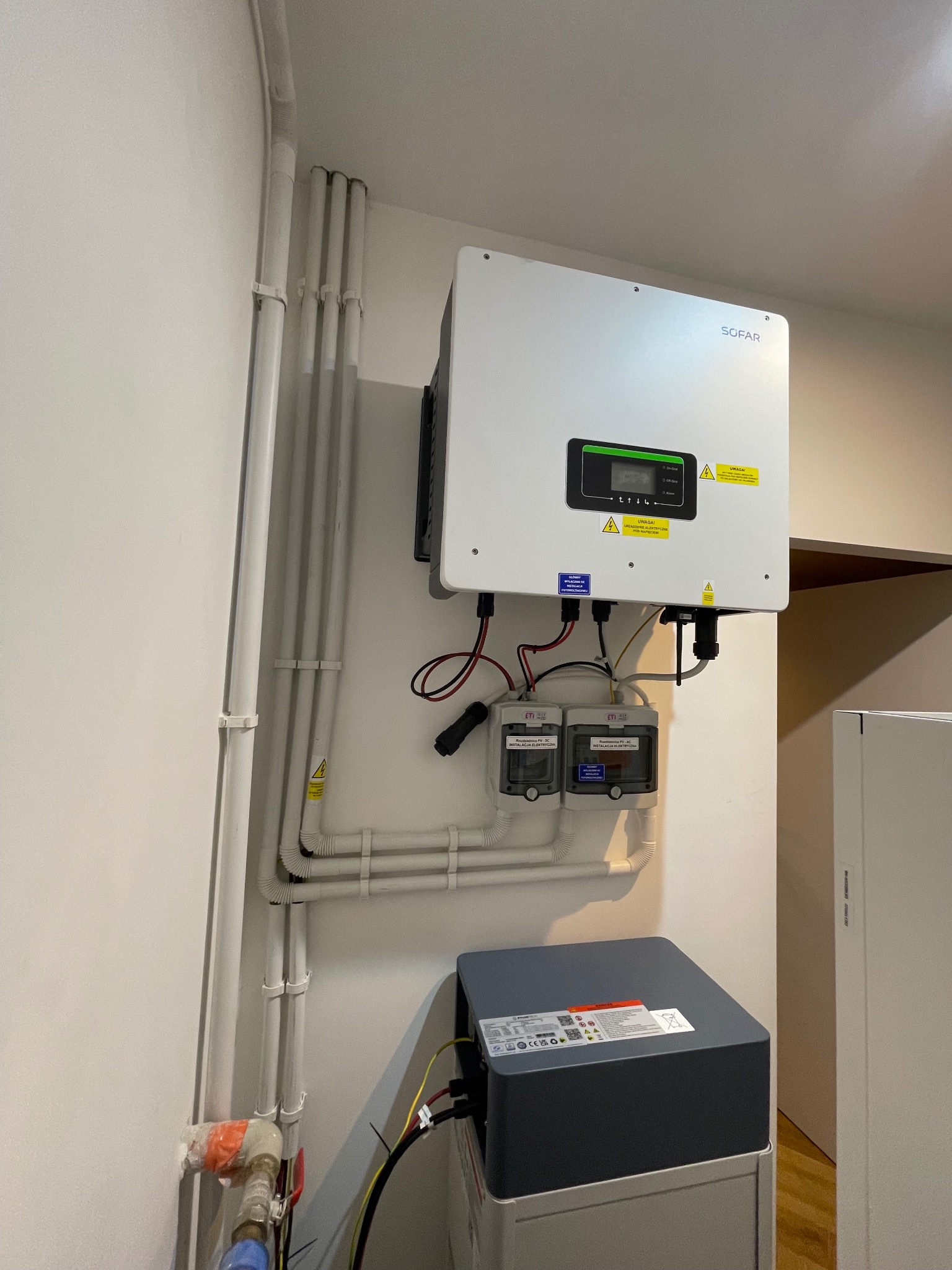 Zamontowany biały falownik Sofar z podłączonymi przewodami, umieszczony na ścianie obok białych rur instalacyjnych i szarego magazynu energii, widoczne zabezpieczenia elektryczne.
