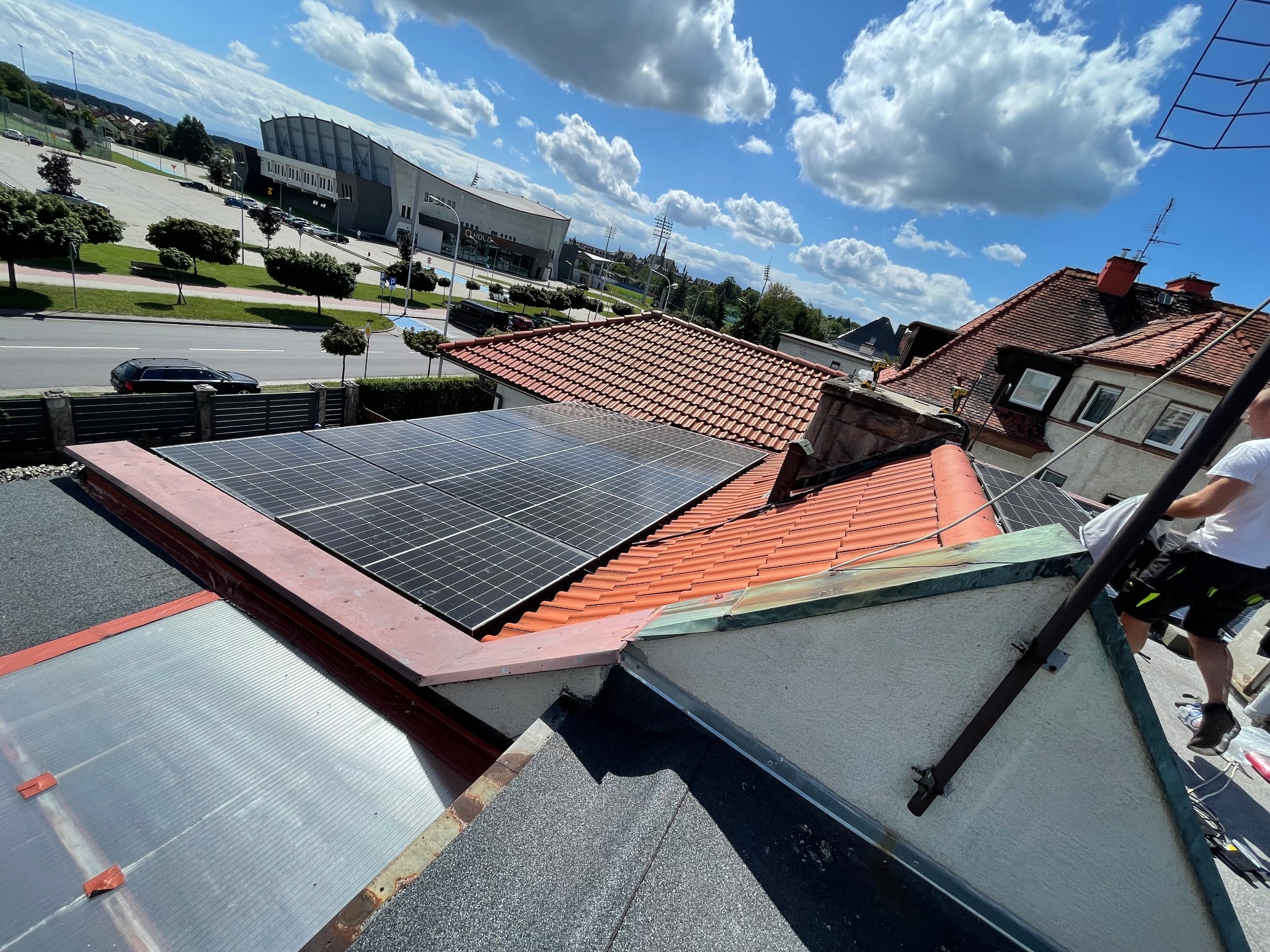 Panele słoneczne zamontowane na czerwonym dachu domu jednorodzinnego, widok z góry, w tle błękitne niebo z chmurami i budynek hali sportowej.