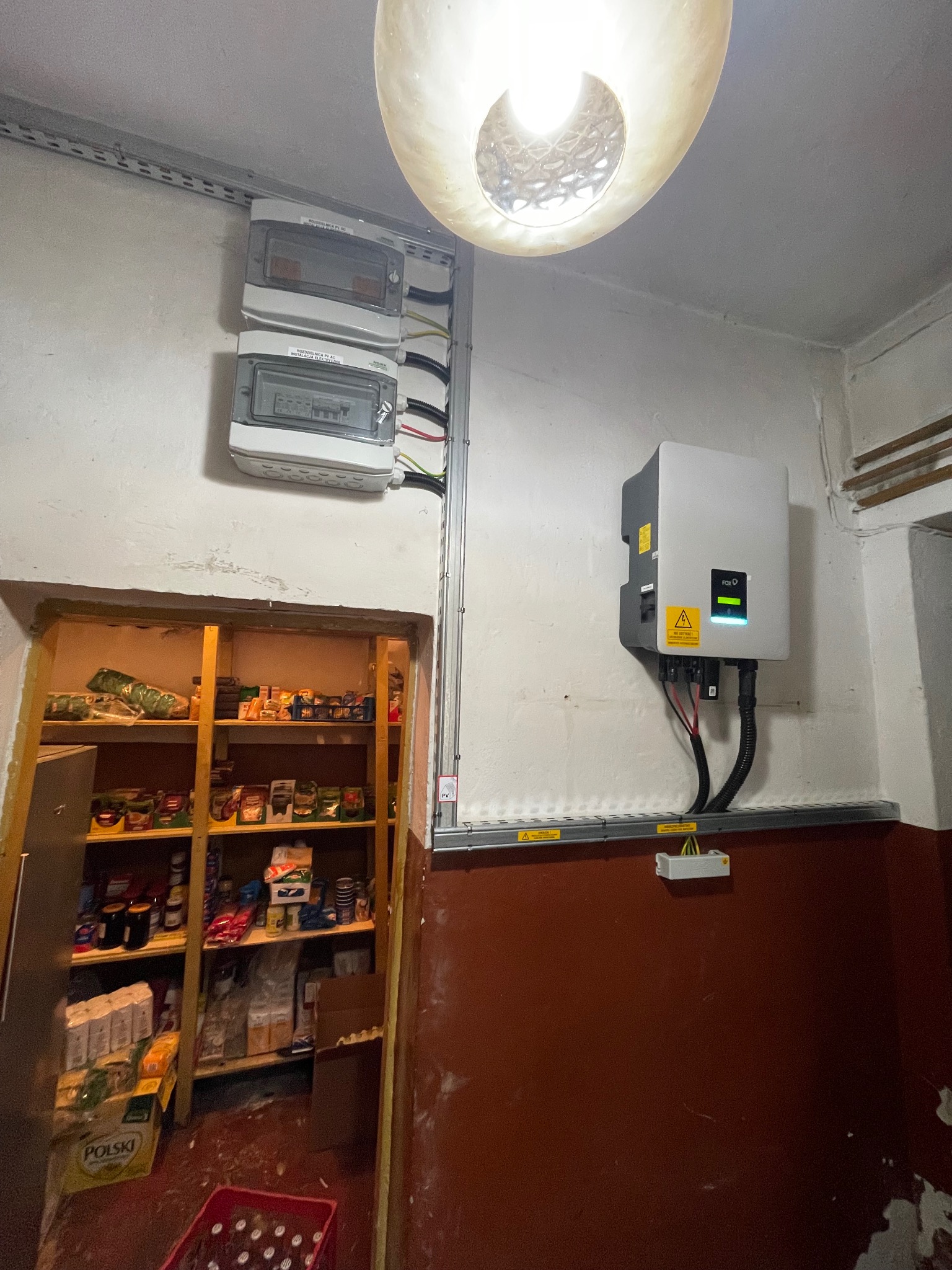 Wnętrze pomieszczenia gospodarczego z zamontowanym inwerterem fotowoltaicznym na ścianie, widoczne skrzynki elektryczne, kable w korytkach oraz regał z zapasami żywności.