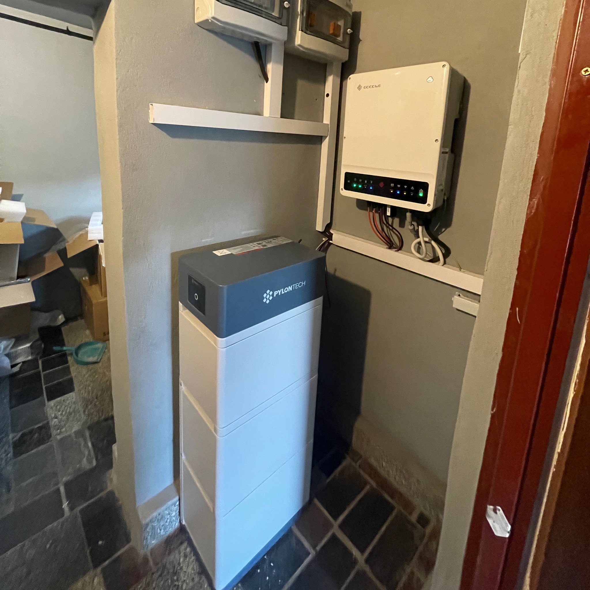 Biały magazyn energii Pylontech z szarym blatem, zamontowany w rogu pomieszczenia z szarą ścianą i brązowymi drzwiami, obok widać biały inwerter z podłączonymi kablami.