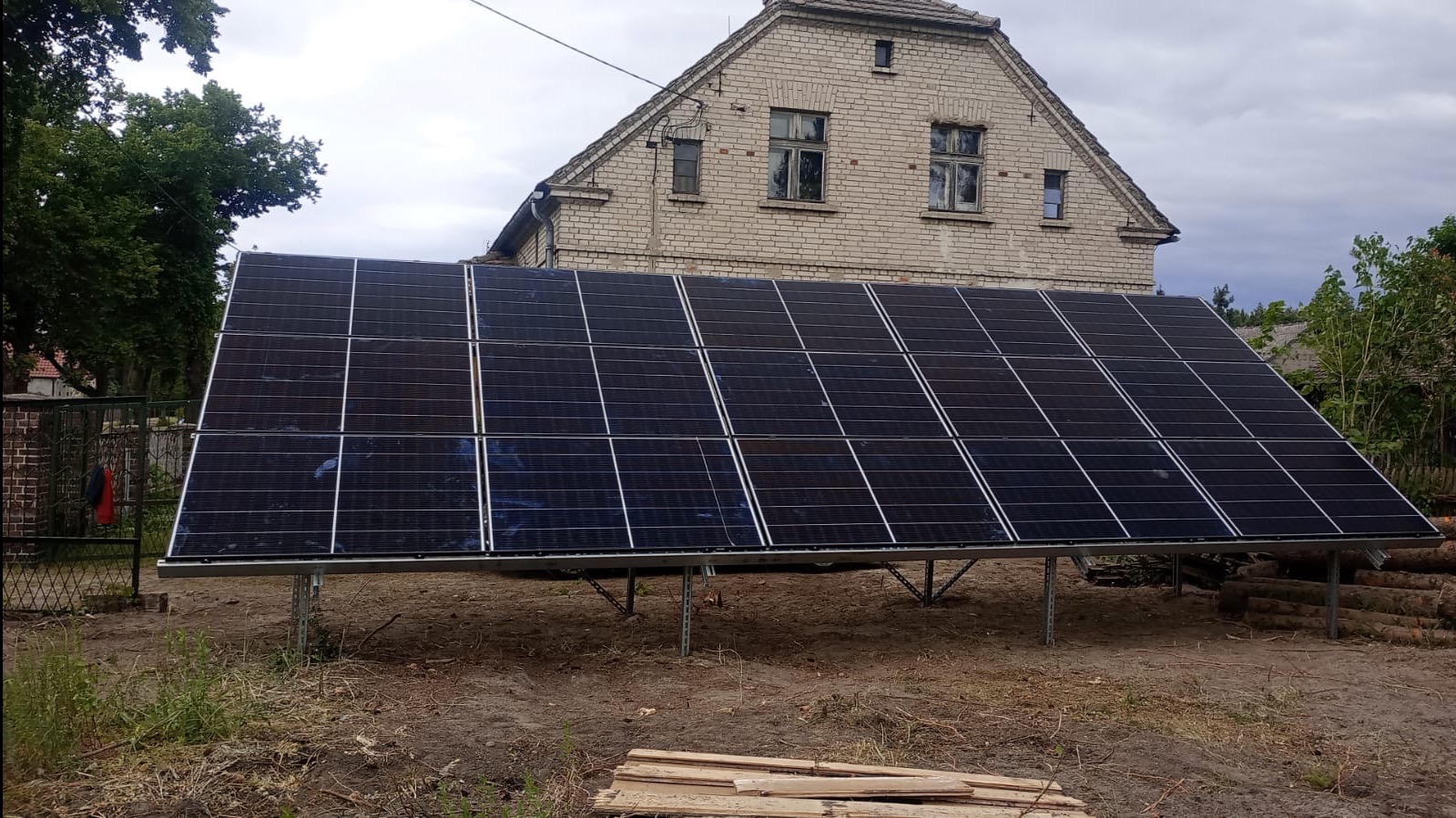 Panel słoneczny ustawiony na ziemi przed ceglanym domem, widoczne drewniane bale i deski na ziemi.