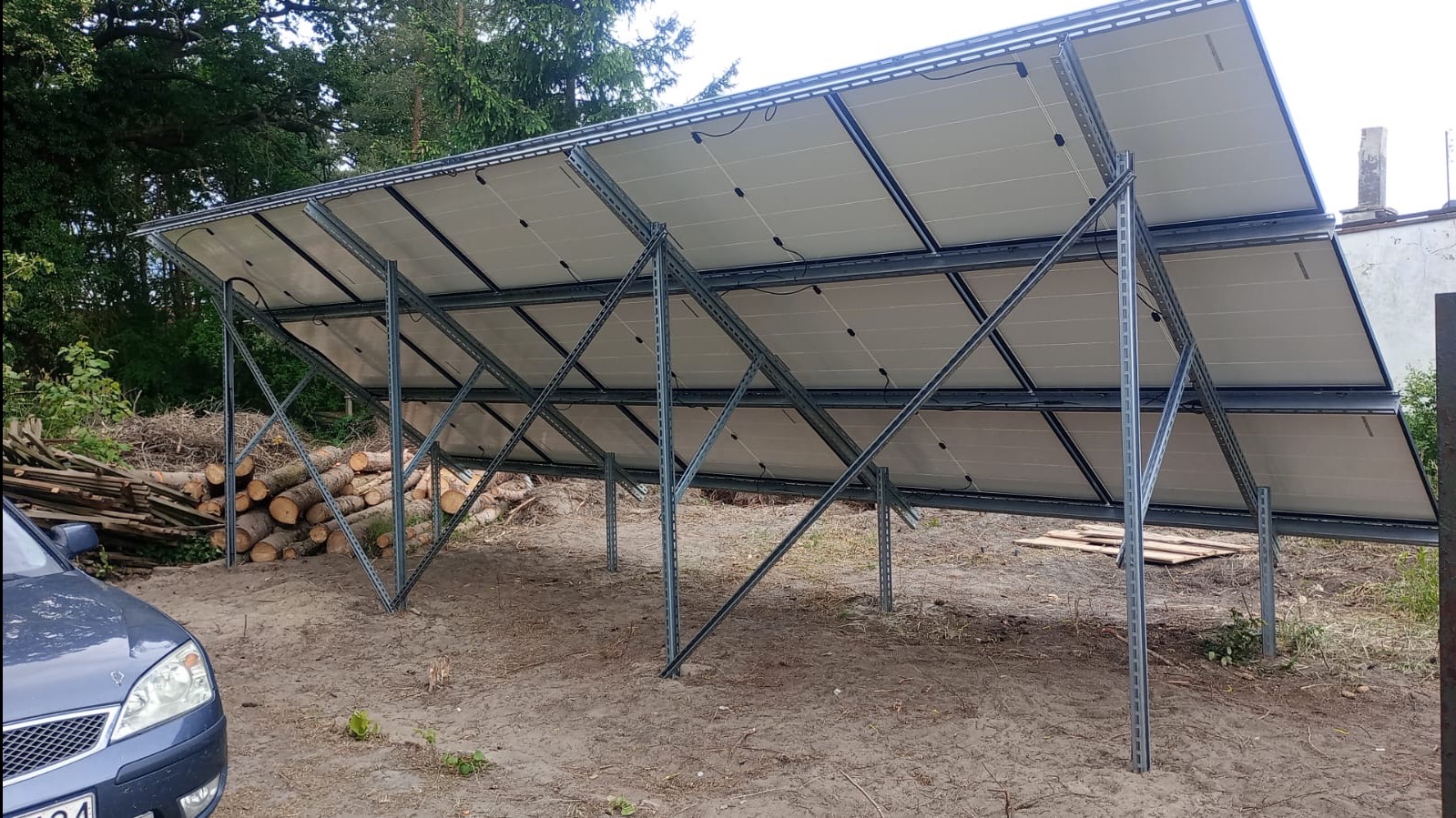 Panele słoneczne zamontowane na metalowej konstrukcji, widok od spodu, z widocznymi kablami i elementami mocującymi, na ziemi leżą pocięte pnie drzew, w tle drzewa i fragment samochodu.