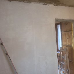 Rafał Aniszewski - Ściana po nałożeniu gładzi, z widocznymi śladami szpachlowania i niedoskonałościami, drabina oparta o ścianę, widok na drewniane szafki przez otwór drzwiowy.