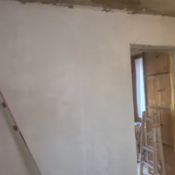 Ściana po nałożeniu gładzi, z widocznymi śladami szpachlowania i niedoskonałościami, drabina oparta o ścianę, widok na drewniane szafki przez otwór drzwiowy.