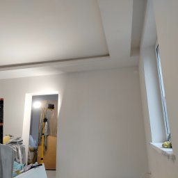 Rafał Aniszewski - Wnętrze w trakcie prac wykończeniowych: świeżo pomalowane ściany na biało, sufit z dekoracyjnym wgłębieniem, okno z zabezpieczoną folią ramą, widoczne narzędzia i lampa budowlana w tle.