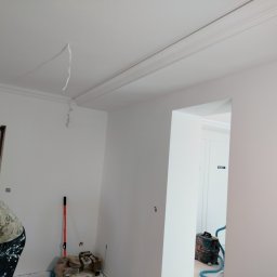 Rafał Aniszewski - Wnętrze w trakcie odświeżania: świeżo pomalowane ściany na biało, widoczne listwy ozdobne pod sufitem, malarz w ubraniu roboczym z plamami farby oraz sprzęt malarski i odkurzacz. Wiszący kabel...