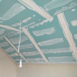 Rafał Aniszewski - Poddasze z zielonymi płytami gips-karton, częściowo pokrytymi białymi paskami szpachli. Widoczna lampa na kablu. Ukośna ściana.