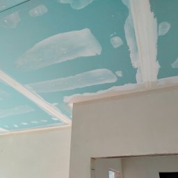 Rafał Aniszewski - Niebieski sufit z płyt kartonowo-gipsowych w trakcie szpachlowania, widoczne łączenia i białe pasy szpachli. Ściany w jasnym odcieniu, fragment otworu drzwiowego.