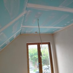 Rafał Aniszewski - Poddasze z zamontowaną płytą karton-gips na skośnym suficie i ścianach, przygotowane do dalszych prac wykończeniowych. Widoczne okno i prowizoryczne oświetlenie.