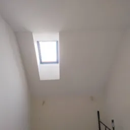 Poddasze po remoncie z oknem dachowym; świeżo pomalowane ściany na biało, widoczne ślady po gładzi, fragment rusztowania w prawym dolnym rogu.