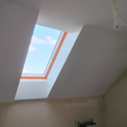 Poddasze z oknem dachowym po remoncie: białe ściany i sufit, widoczne niebo. Częściowo wykończona ściana na dole. Wiszące kable elektryczne.