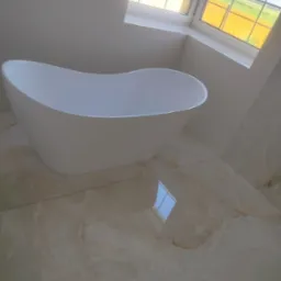 Nowoczesna, wolnostojąca wanna w jasnej łazience z oknem. Beżowe płytki podłogowe z marmurkowym wzorem odbijają światło. Minimalistyczny design.