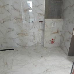 Rafał Aniszewski - Wykończona łazienka z marmurowymi płytkami w odcieniach bieli i beżu. Widoczne przyłącza wodne i odpływ liniowy. Częściowo widoczny mur z cegły.