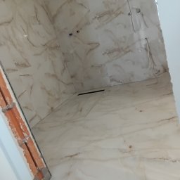 Rafał Aniszewski - Wykończona łazienka z beżowymi płytkami imitującymi marmur na ścianach i podłodze. Widoczne instalacje wodne i fragment niedokończonego obramowania drzwi.