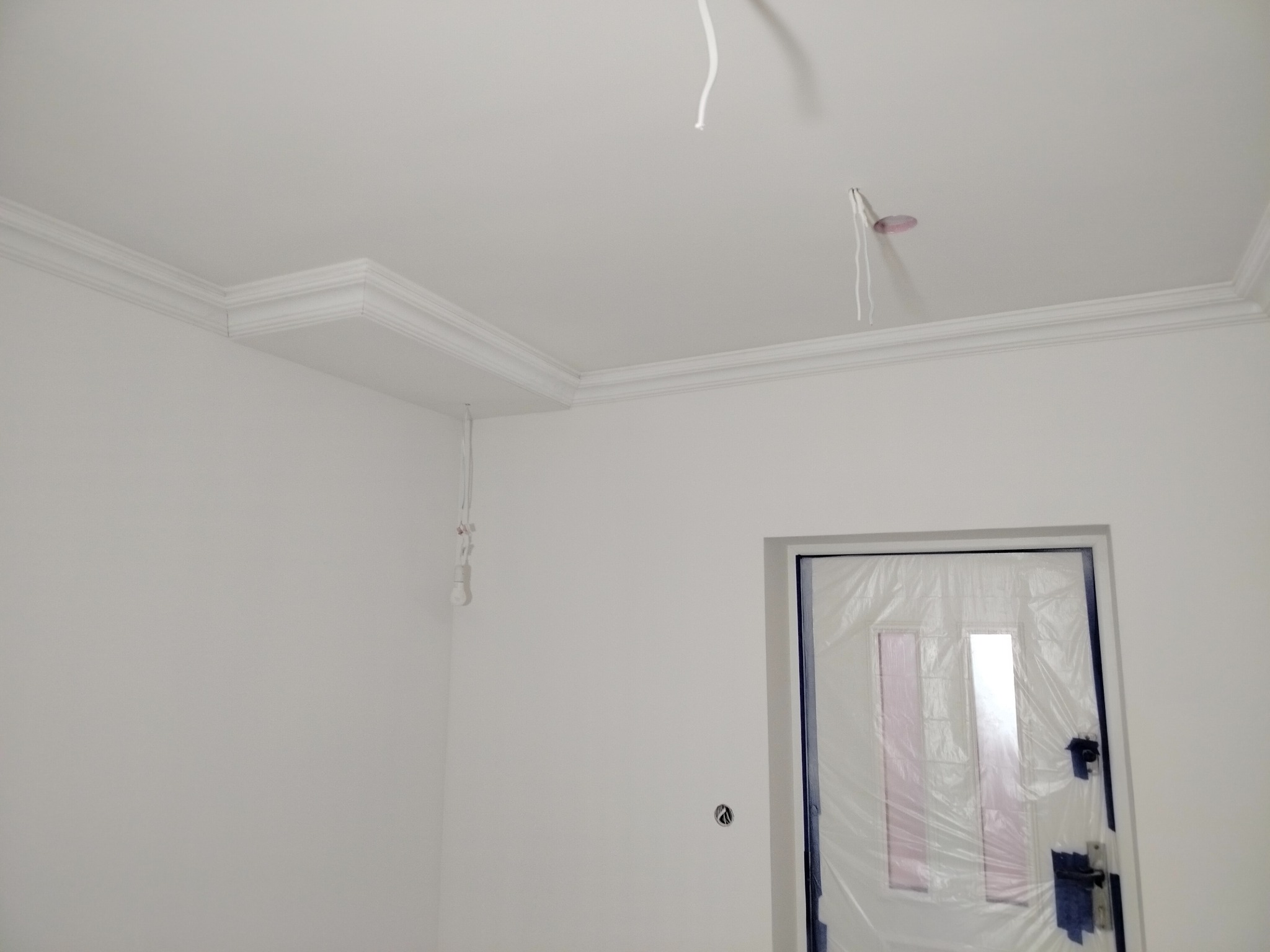 Białe wnętrze w trakcie wykańczania: świeżo pomalowane ściany i sufit z dekoracyjną sztukaterią, zabezpieczone drzwi folią ochronną, wystające przewody elektryczne.