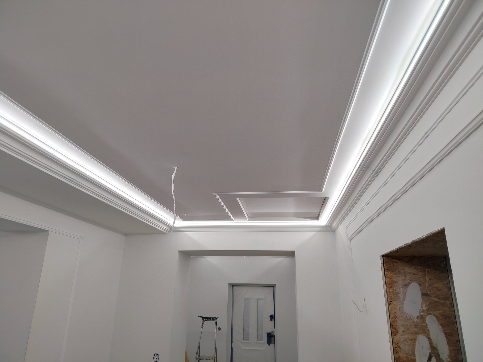 Biały sufit z dekoracyjnymi listwami LED i gładkimi ścianami w trakcie wykańczania, widoczna drabina i zabezpieczone drzwi.
