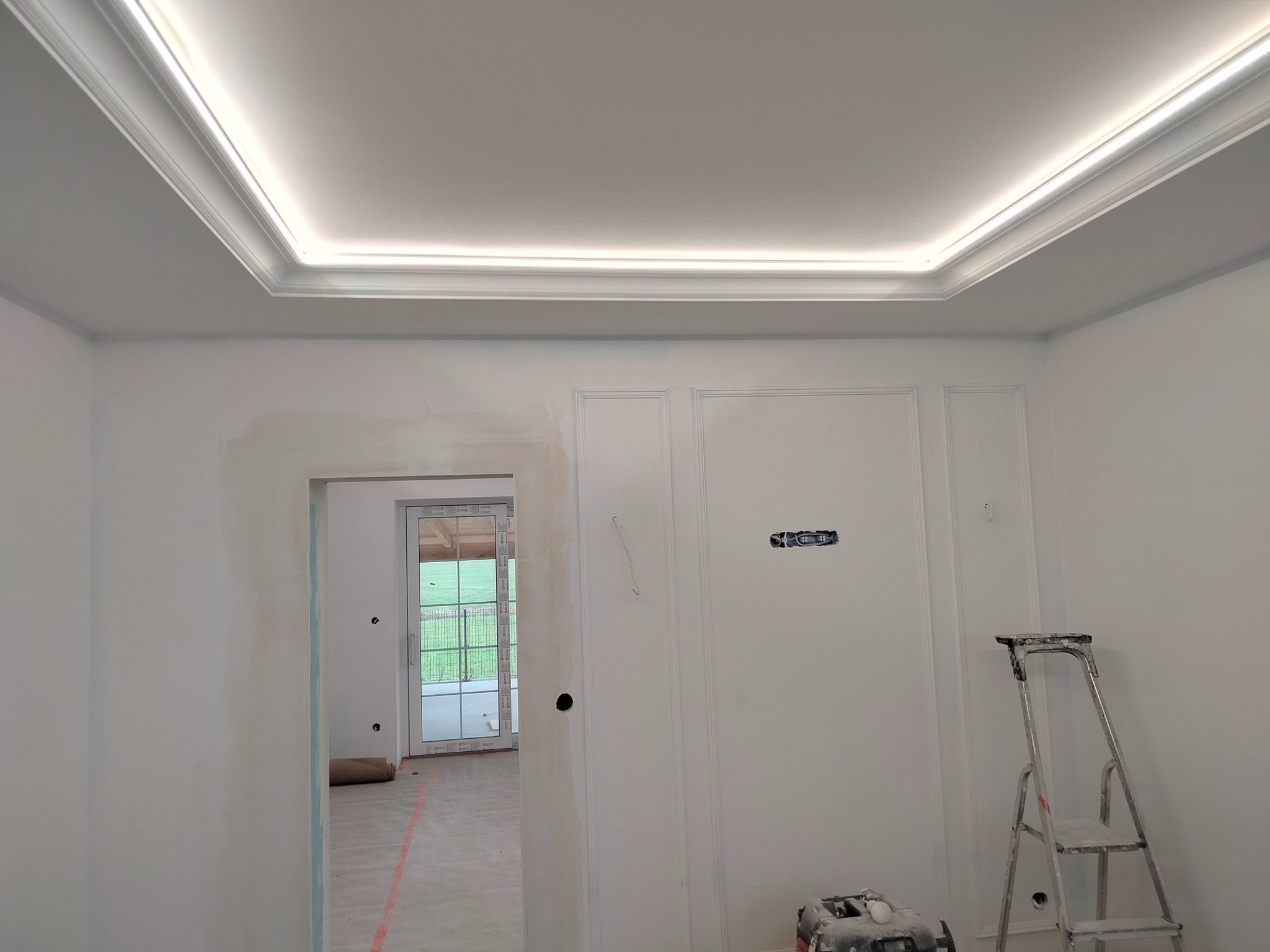 Wnętrze w trakcie prac wykończeniowych: świeżo pomalowane ściany z dekoracyjnymi panelami, podświetlany sufit z listwami, drabina malarska i widoczny otwór drzwiowy prowadzący na zewnątrz.