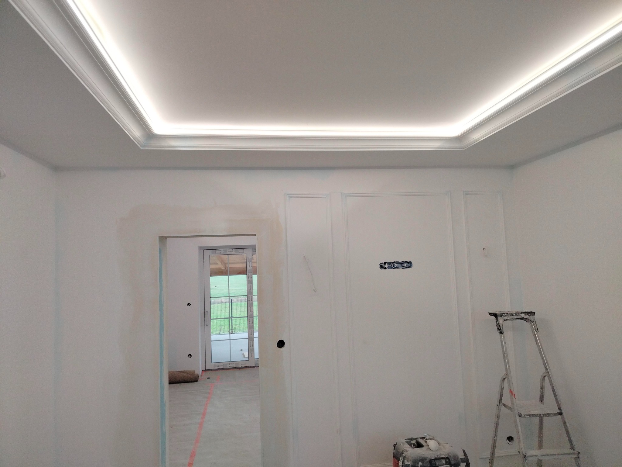 Wnętrze w trakcie prac wykończeniowych: widoczne ślady po szpachlowaniu na ścianie, sufit z dekoracyjnym oświetleniem LED, drabina malarska i skrzynka narzędziowa.