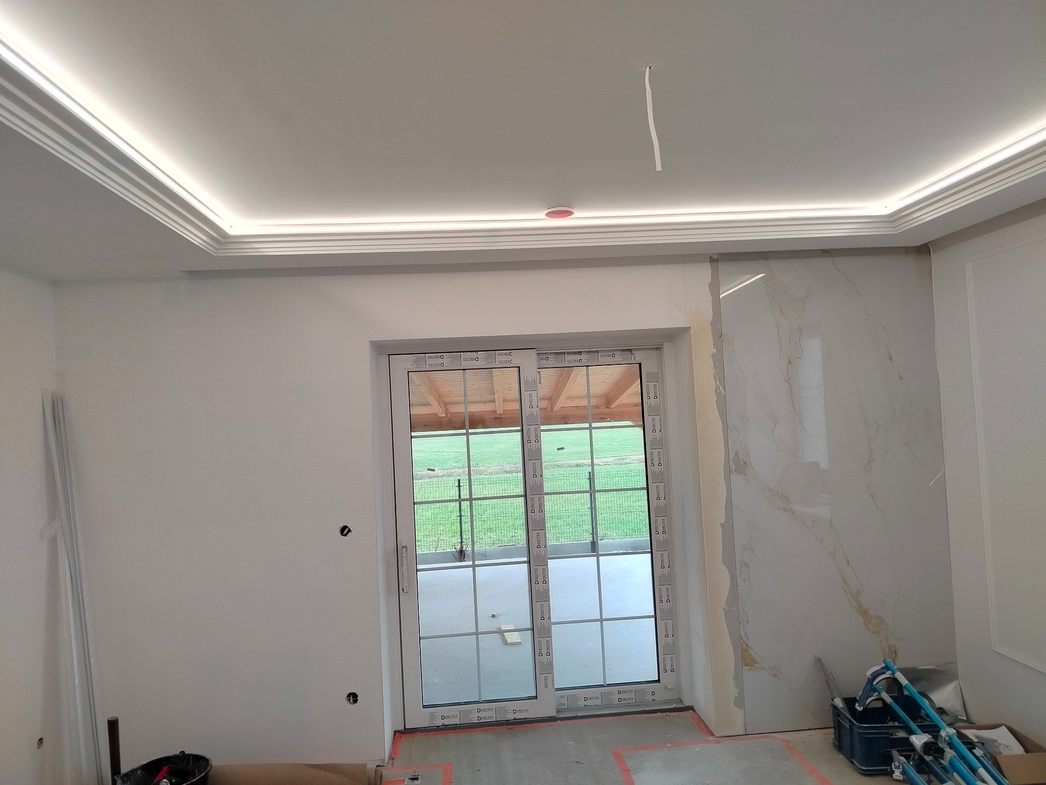 Wnętrze w trakcie wykańczania: sufit z dekoracyjną listwą LED, fragment ściany z płytą imitującą marmur, widoczne narzędzia i materiały budowlane, okno balkonowe z widokiem na zieleń.