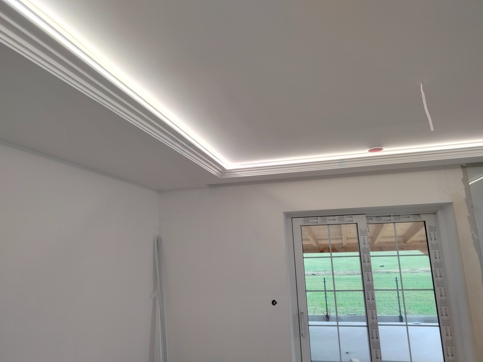 Nowoczesne wykończenie sufitu z listwami LED, widok na okno przesuwne z widokiem na zewnątrz, ściany przygotowane do malowania.