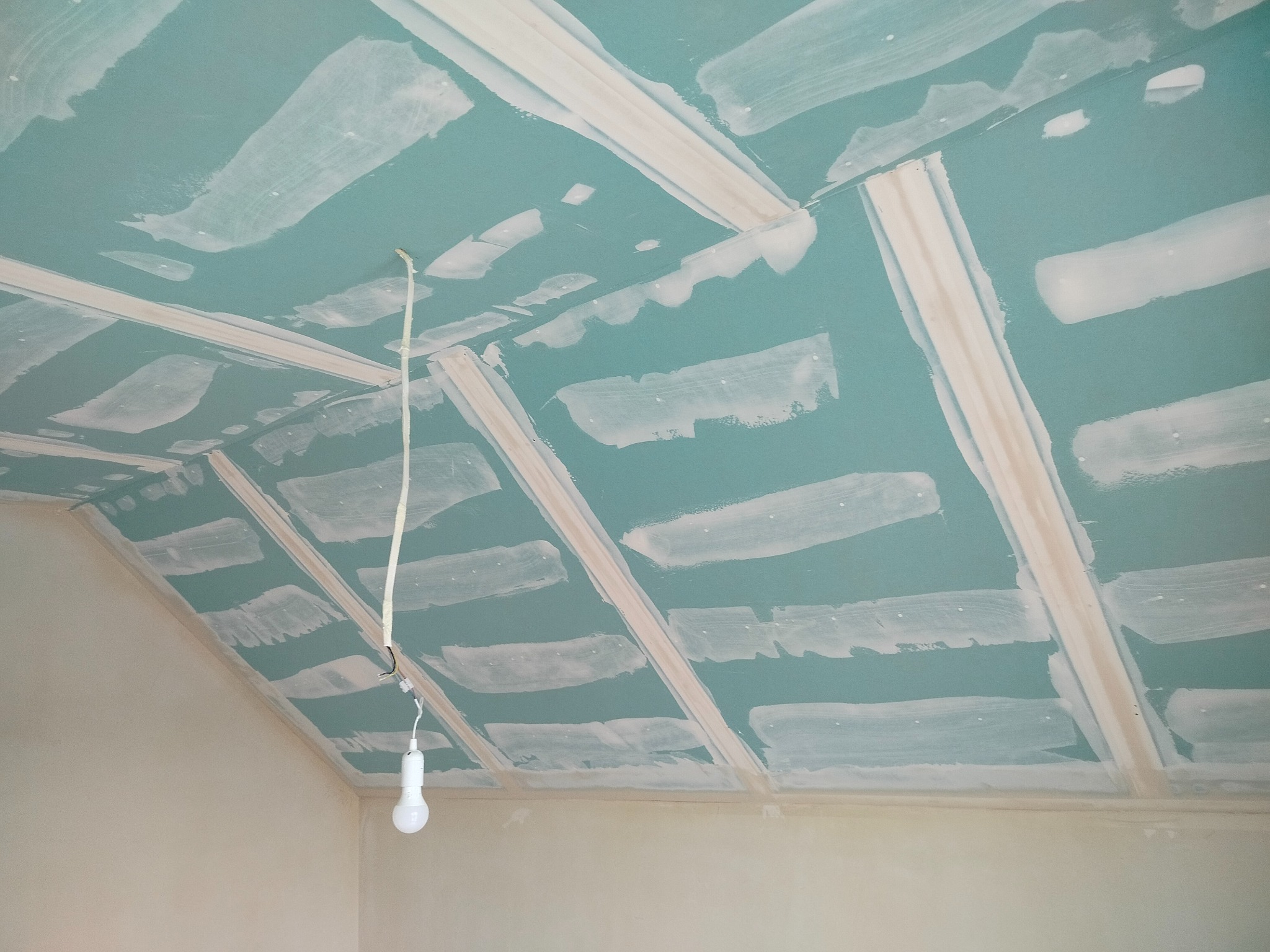 Poddasze z zielonymi płytami gips-karton, częściowo pokrytymi białymi paskami szpachli. Widoczna lampa na kablu. Ukośna ściana.