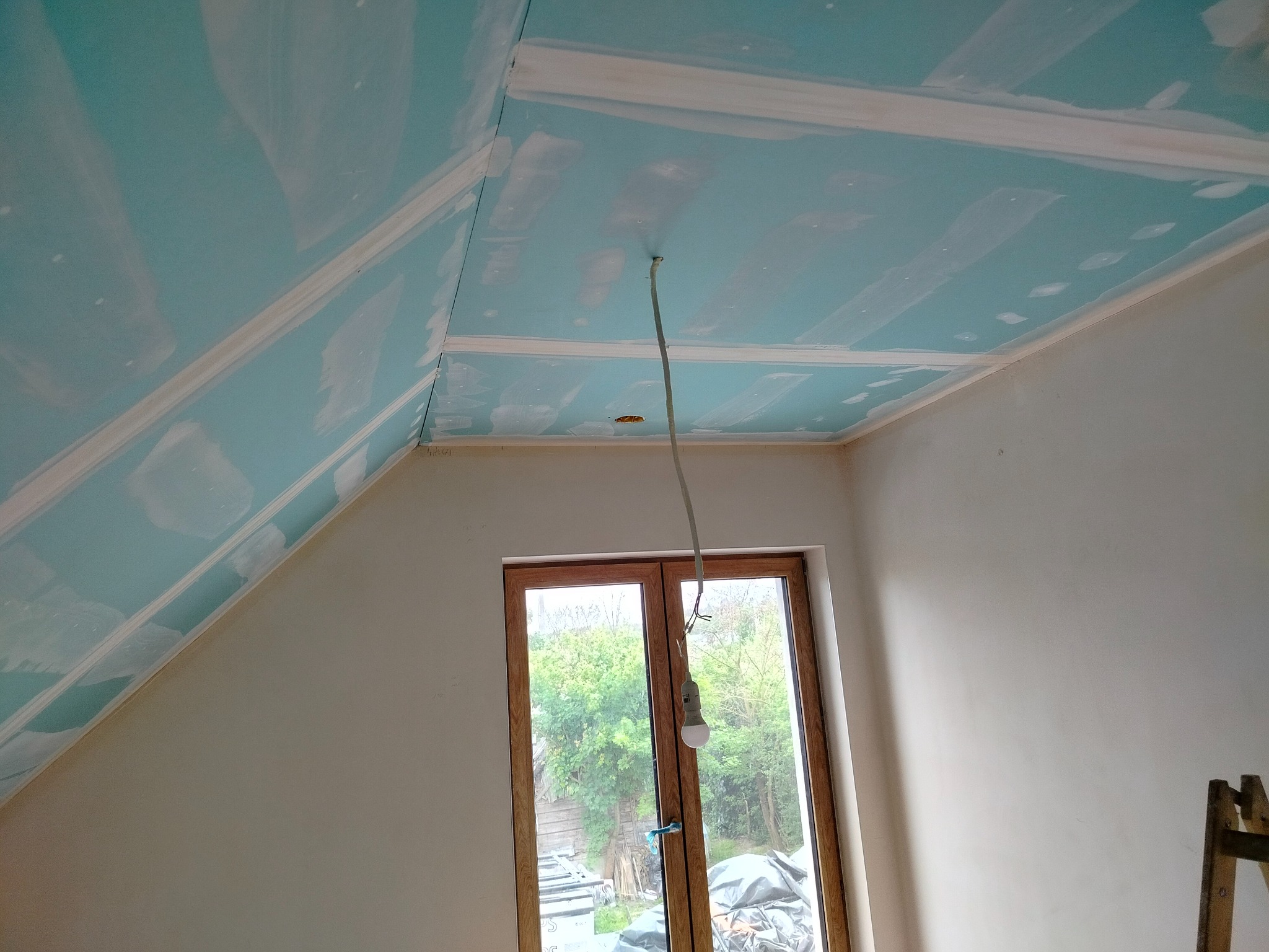 Poddasze z zamontowaną płytą karton-gips na skośnym suficie i ścianach, przygotowane do dalszych prac wykończeniowych. Widoczne okno i prowizoryczne oświetlenie.