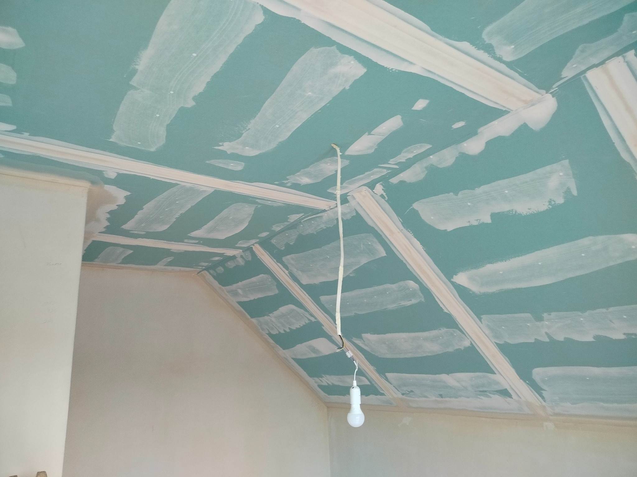 Skośny sufit z płyt kartonowo-gipsowych, wstępnie wyszpachlowany, z widoczną konstrukcją szkieletową i wiszącą żarówką, przygotowany do dalszych prac wykończeniowych.