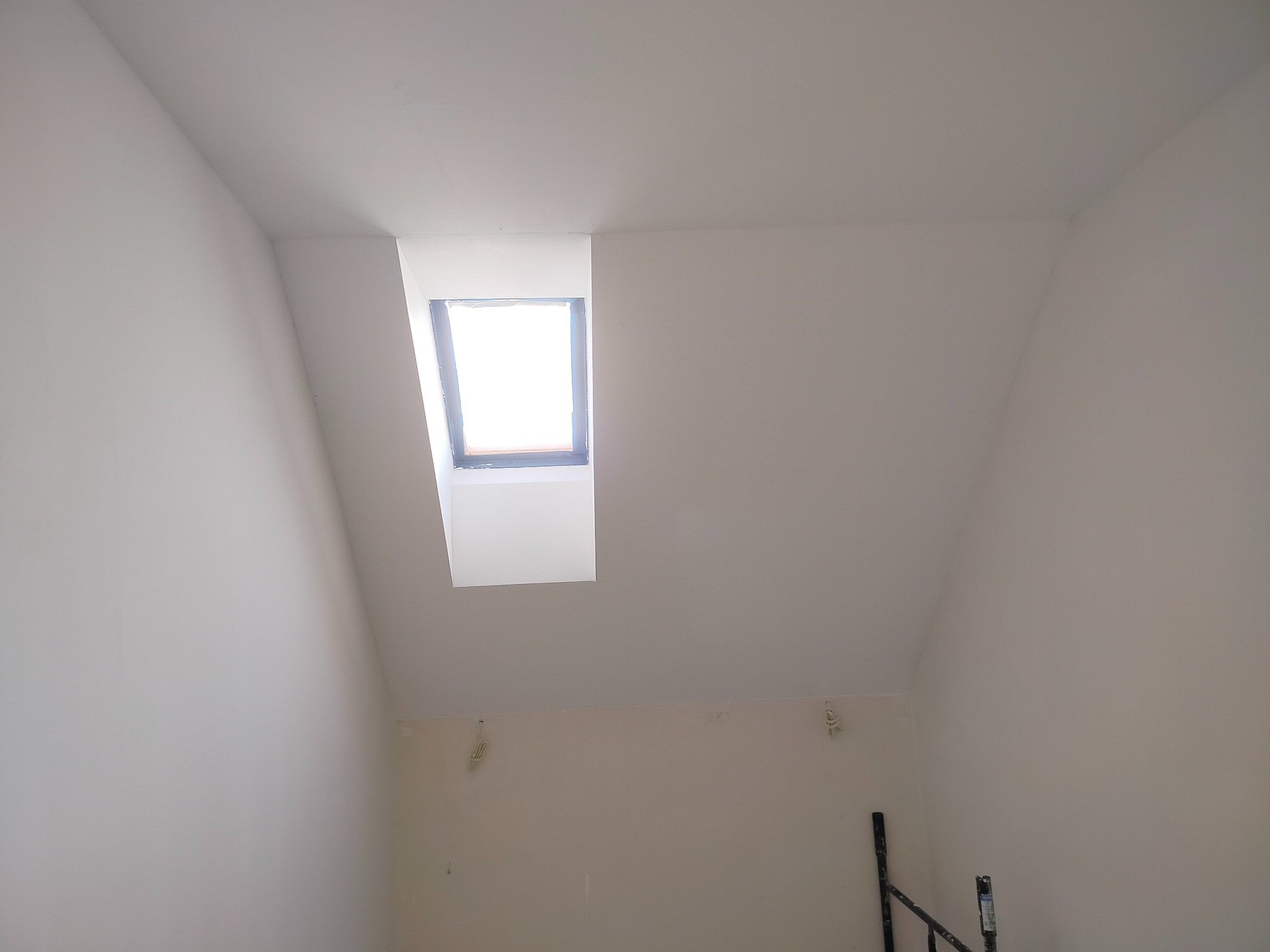 Poddasze po remoncie z oknem dachowym; świeżo pomalowane ściany na biało, widoczne ślady po gładzi, fragment rusztowania w prawym dolnym rogu.