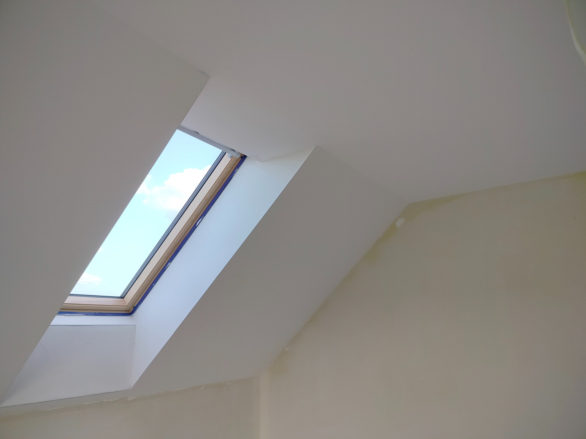 Poddasze po szpachlowaniu i malowaniu na biało, widoczne okno dachowe z niebieskim niebem i chmurami. Ściany i sufit gładkie, przygotowane do dalszych prac.