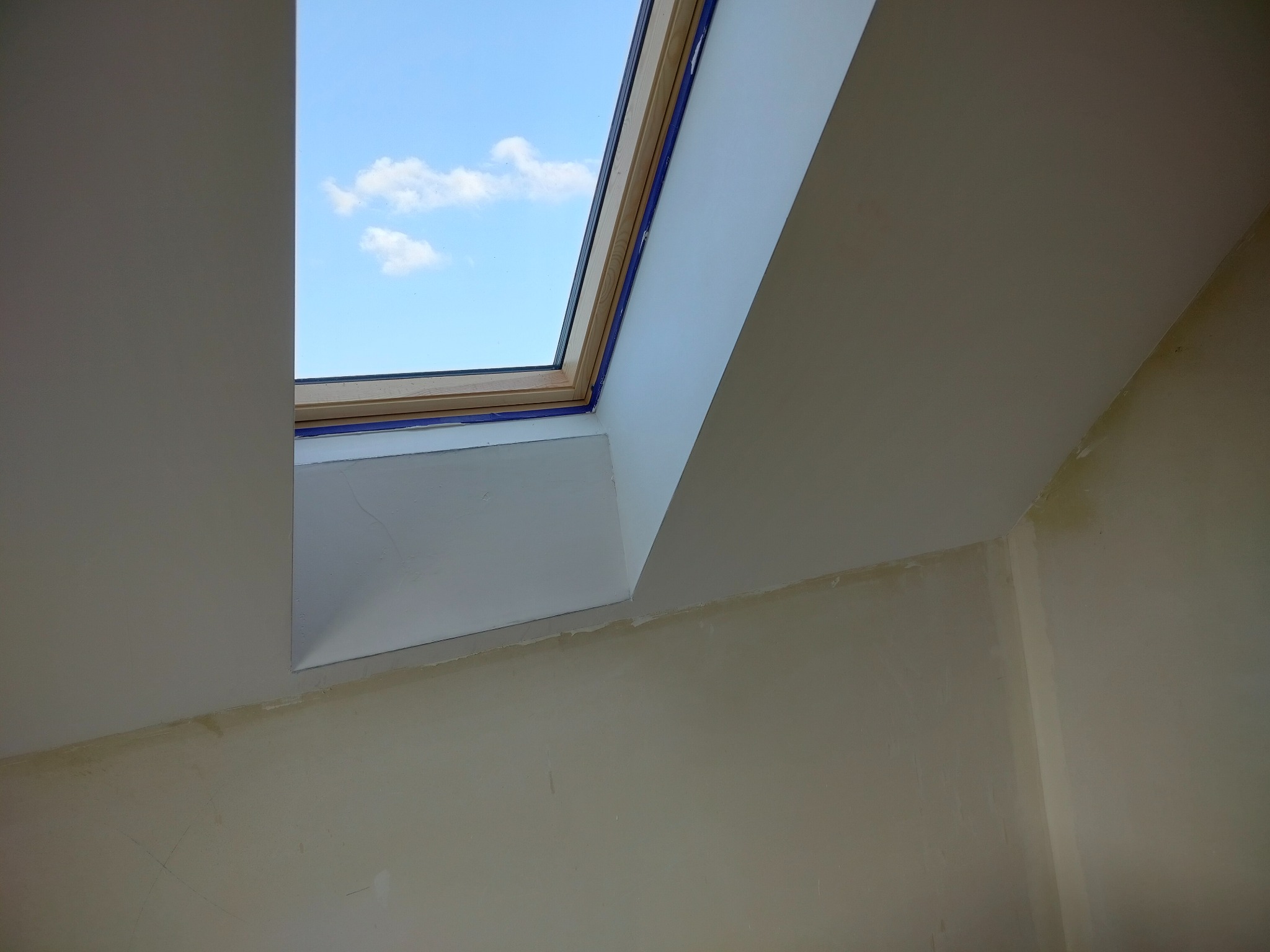 Poddasze z oknem dachowym w trakcie prac wykończeniowych. Widoczne zabezpieczenia taśmą malarską i surowa ściana przed malowaniem.