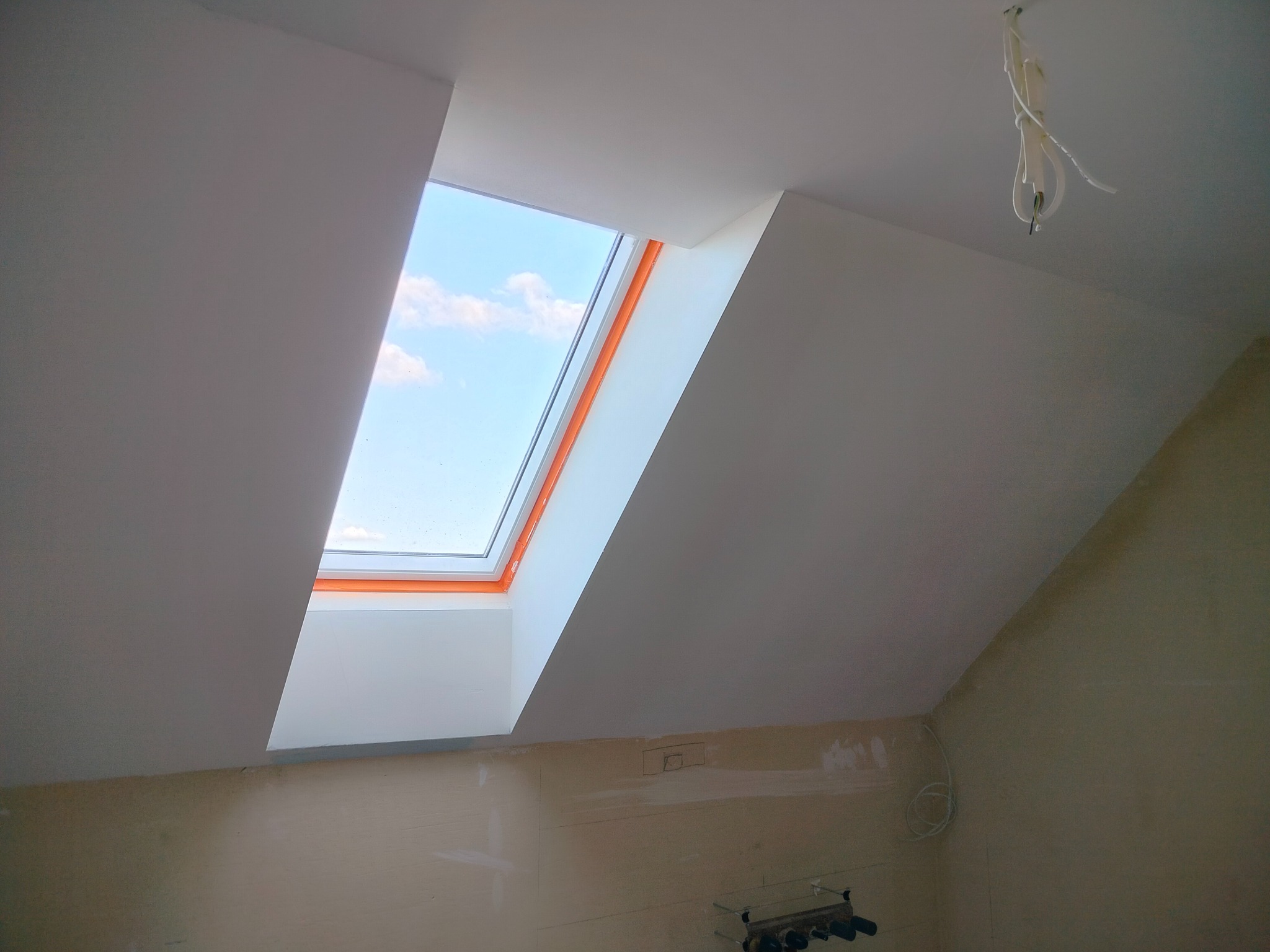 Poddasze z oknem dachowym po remoncie: białe ściany i sufit, widoczne niebo. Częściowo wykończona ściana na dole. Wiszące kable elektryczne.