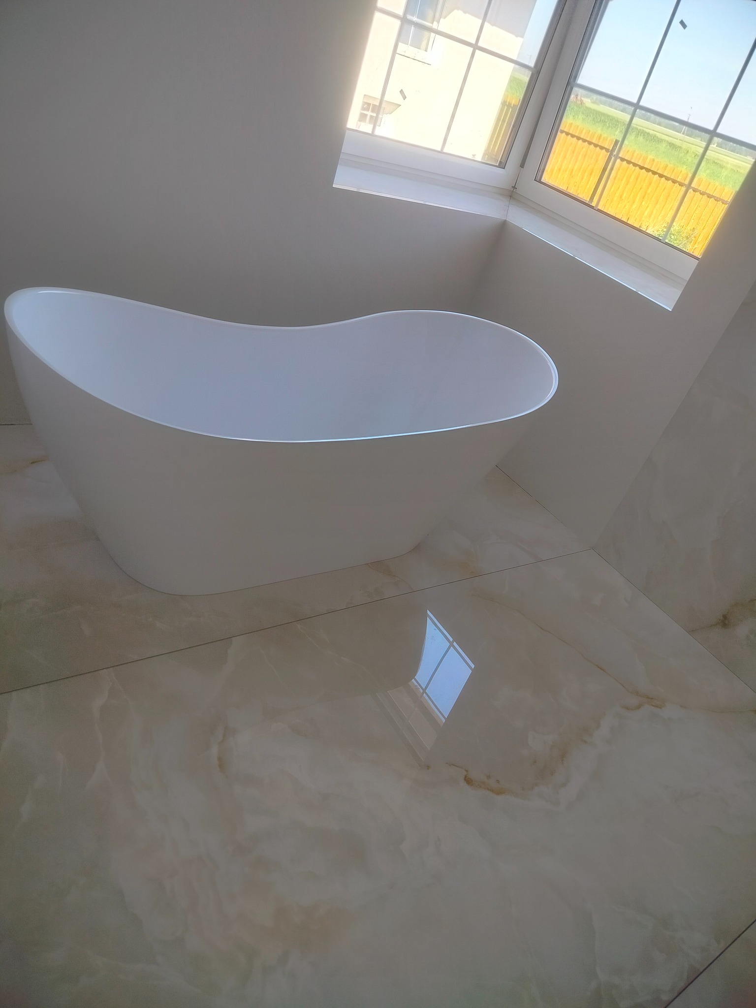 Nowoczesna, wolnostojąca wanna w jasnej łazience z oknem. Beżowe płytki podłogowe z marmurkowym wzorem odbijają światło. Minimalistyczny design.