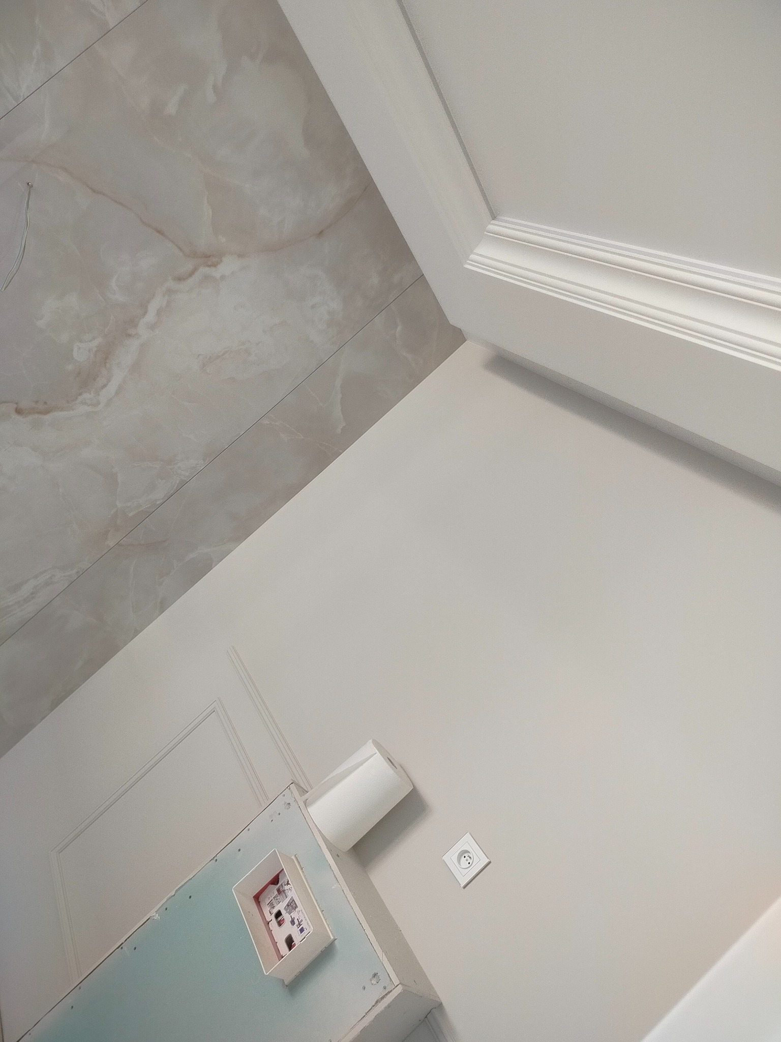 Wykończone wnętrze z marmurową okładziną sufitu, sztukaterią i gładką, pomalowaną ścianą z gniazdkiem elektrycznym. Widoczna płyta karton-gips z puszką instalacyjną.