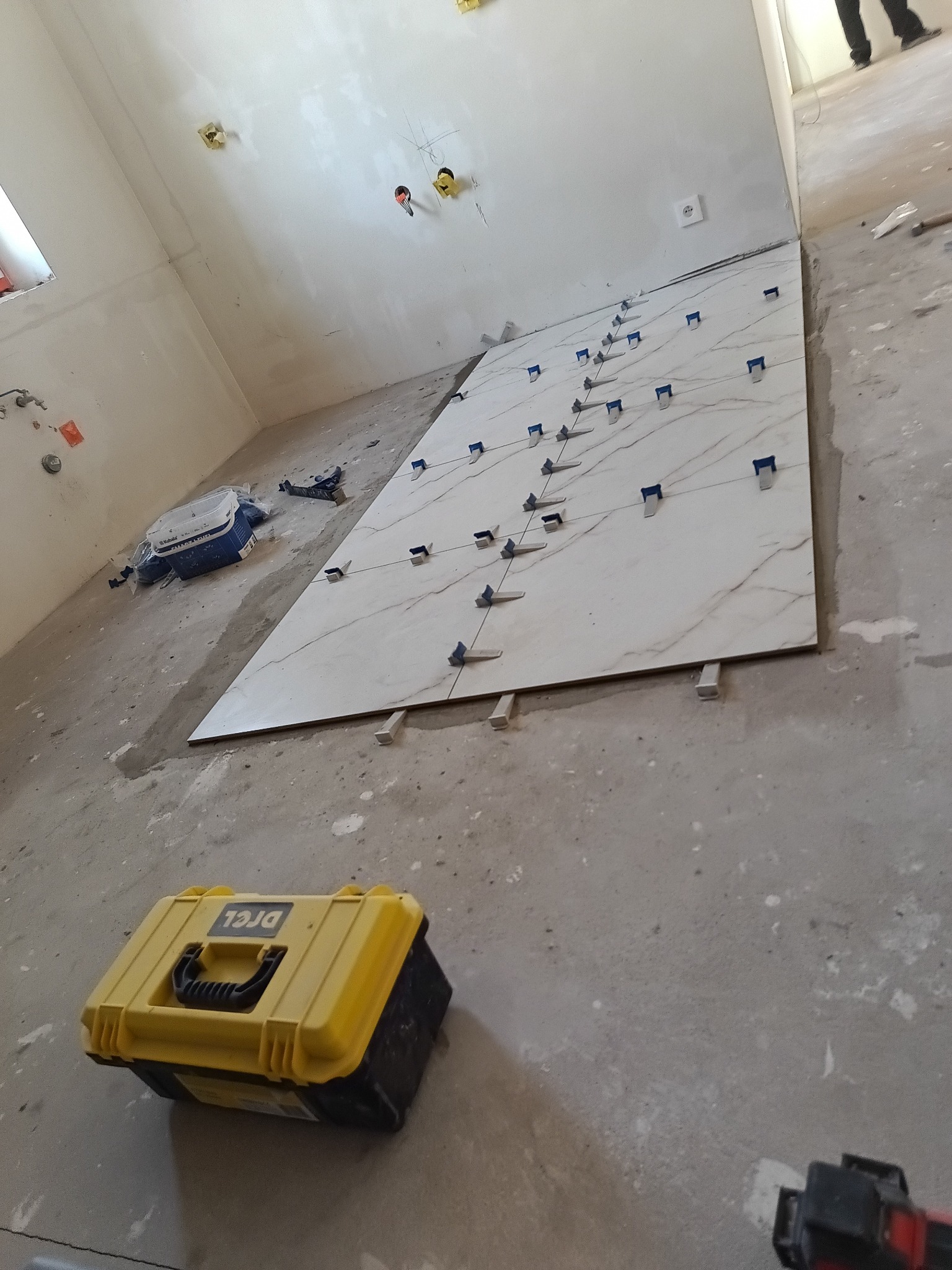 Układanie wielkoformatowych płytek z użyciem systemu poziomowania. Żółta skrzynka narzędziowa w dolnej części kadru. Widoczne instalacje elektryczne w ścianie.