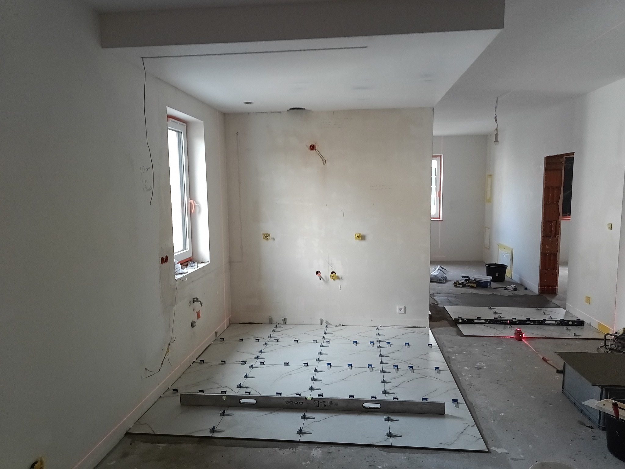 Wnętrze w trakcie układania płytek podłogowych. Widoczne poziomice, krzyżyki dystansowe i laserowy niwelator. Ściany przygotowane do dalszych prac wykończeniowych.