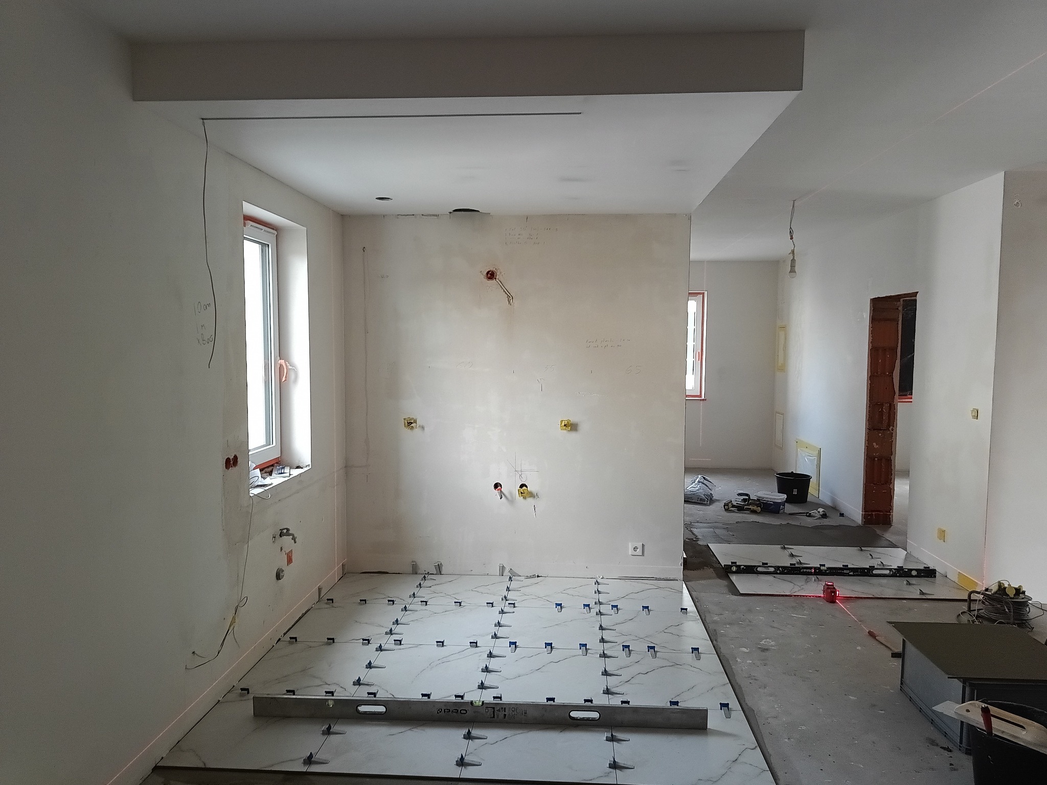 Układanie białych płytek z szarym wzorem w nowym wnętrzu. Widoczne poziomice, krzyżyki dystansowe i instalacje elektryczne. Laserowy niwelator wyznacza poziom.