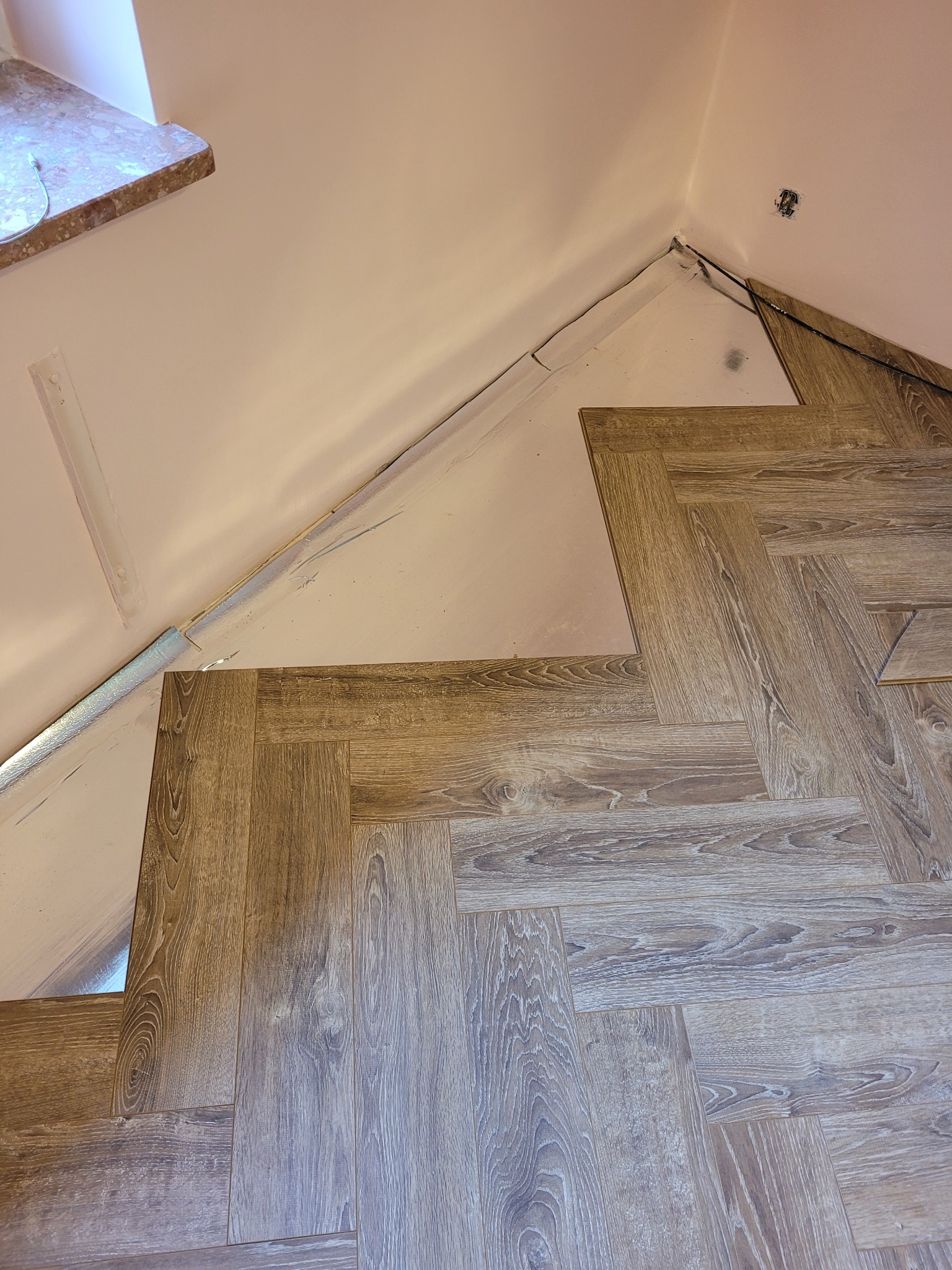 Układanie paneli podłogowych w jodełkę w rogu pomieszczenia, widoczna folia izolacyjna przy ścianie, gniazdko elektryczne na ścianie.