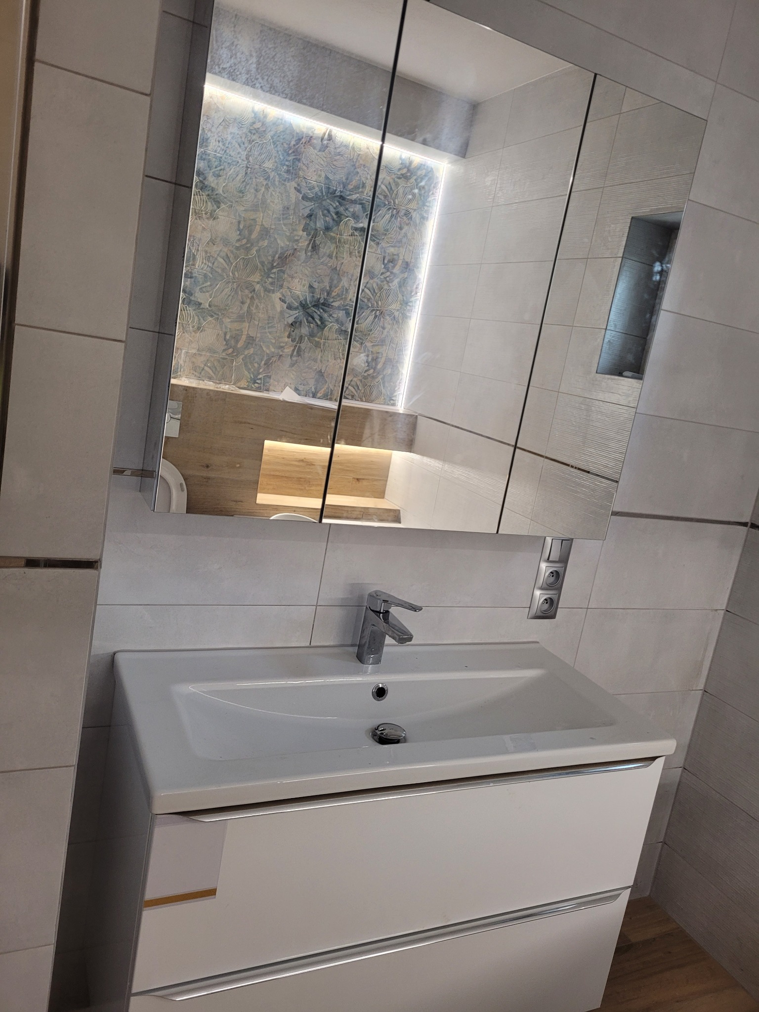 Nowoczesna łazienka po remoncie: biała umywalka z szafką, bateria, lustro z szafką wiszącą odbijające fragment toalety z wzorzystą tapetą i półkami z oświetleniem LED.