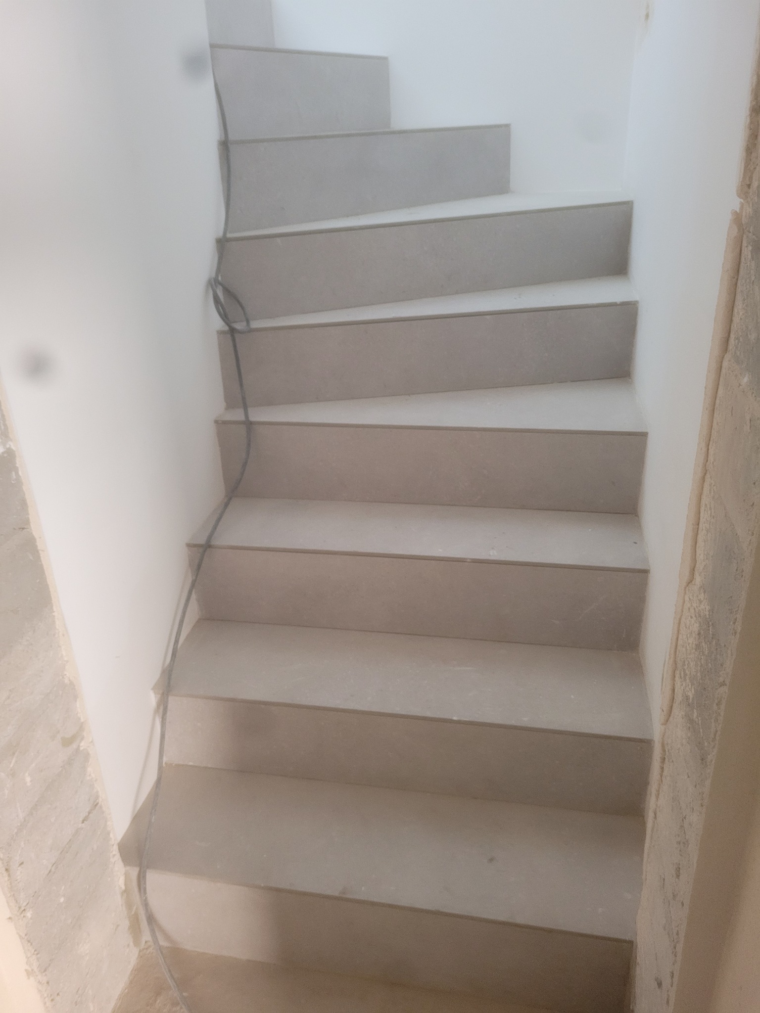 Schody wyłożone szarymi płytkami w trakcie montażu, widoczne niedokończone ściany i kabel zwisający wzdłuż ściany.