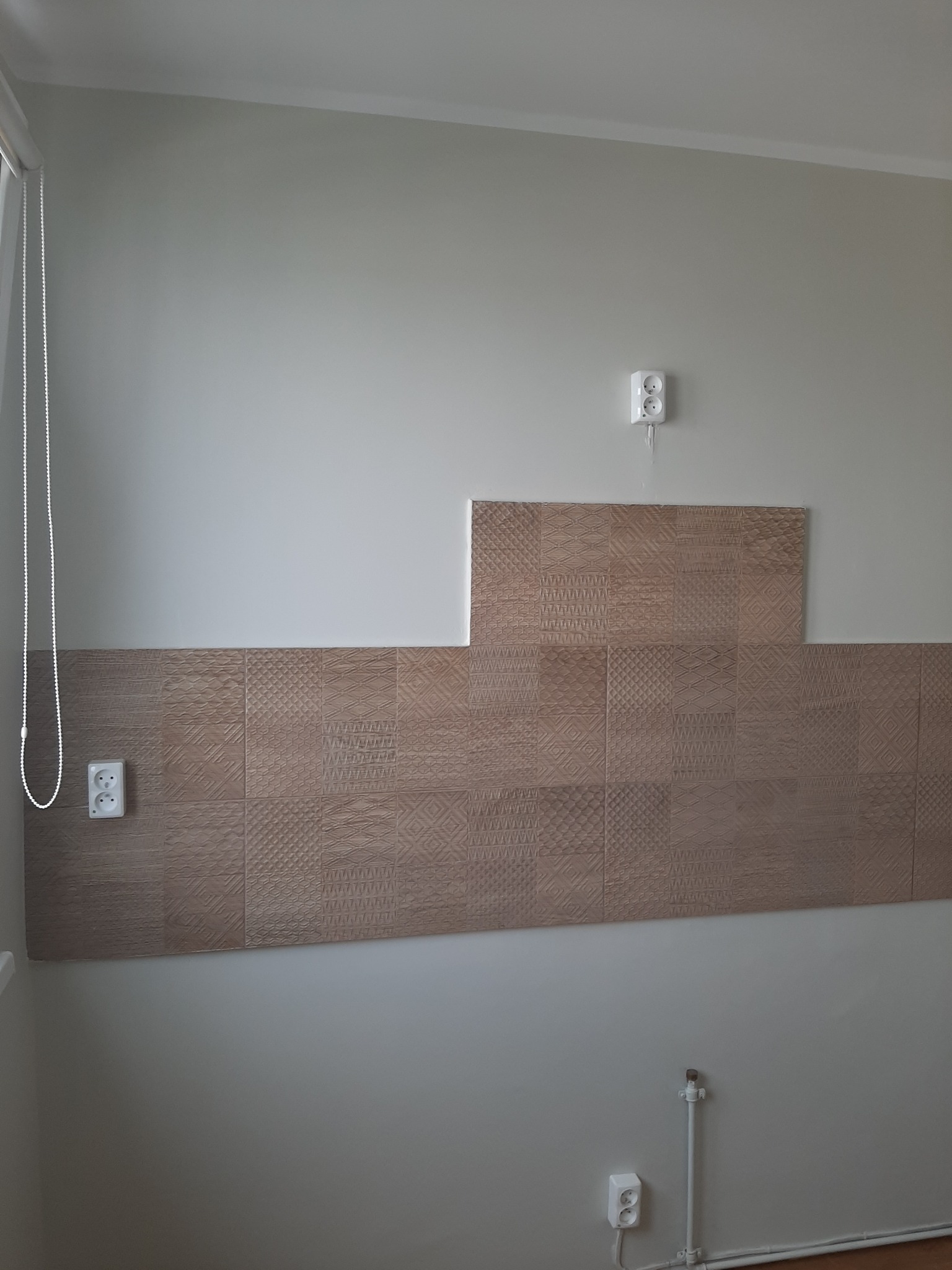 Fragment ściany z częściowo ułożonymi dekoracyjnymi płytkami ceramicznymi w odcieniach brązu, widoczne gniazdka elektryczne i rura doprowadzająca wodę.