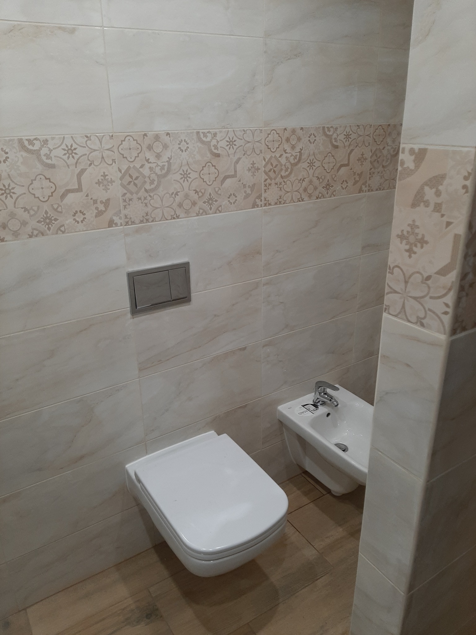 Wykończona łazienka z beżowymi płytkami imitującymi marmur, dekoracyjnym pasem z motywami kwiatowymi i geometrycznymi, wiszącą toaletą oraz bidetem z chromowaną baterią.