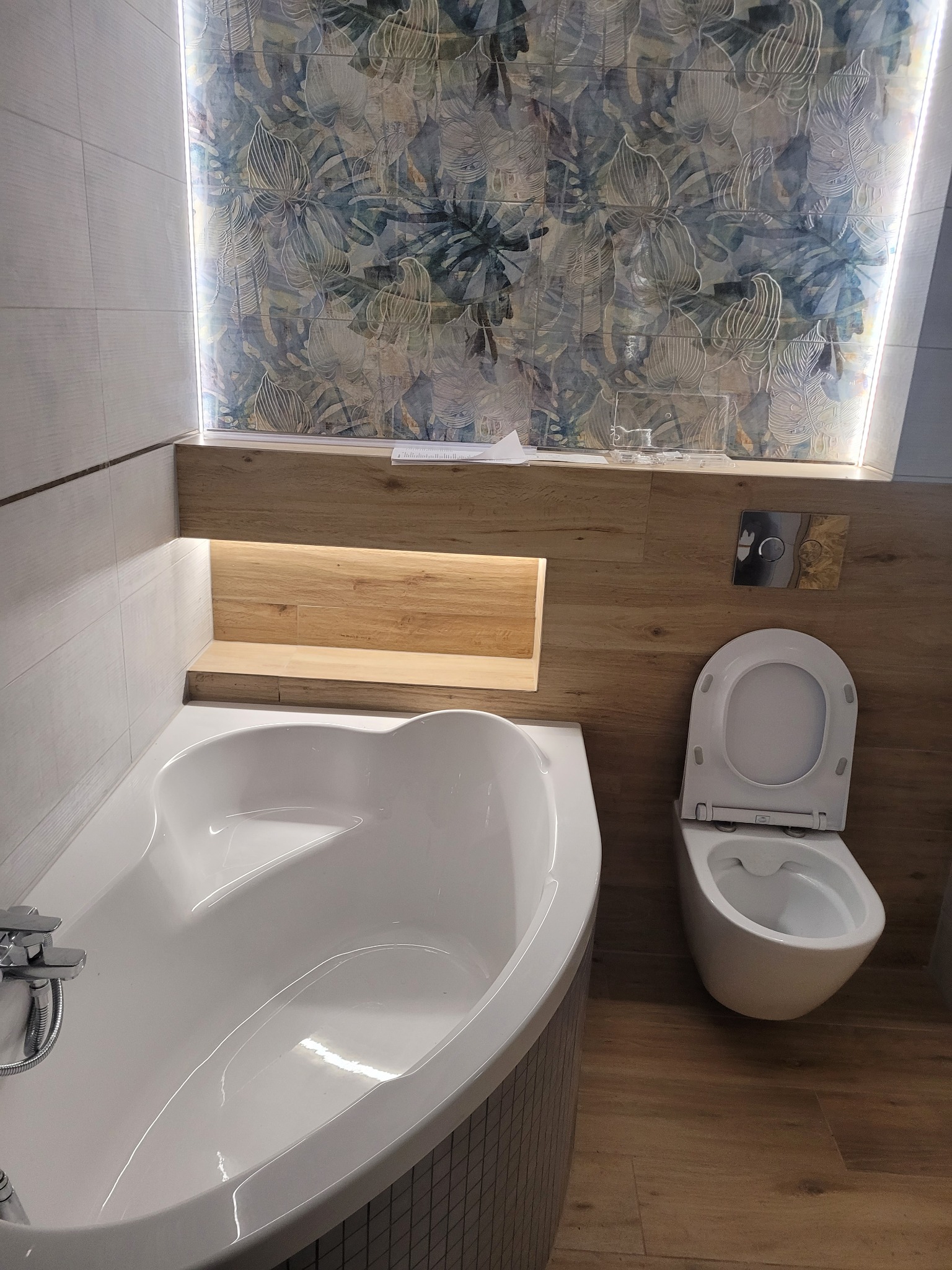 Nowoczesna łazienka z wanną o nietypowym kształcie, wiszącą toaletą i podświetlaną ścianą z motywem roślinnym, wykończona drewnopodobnymi płytkami.