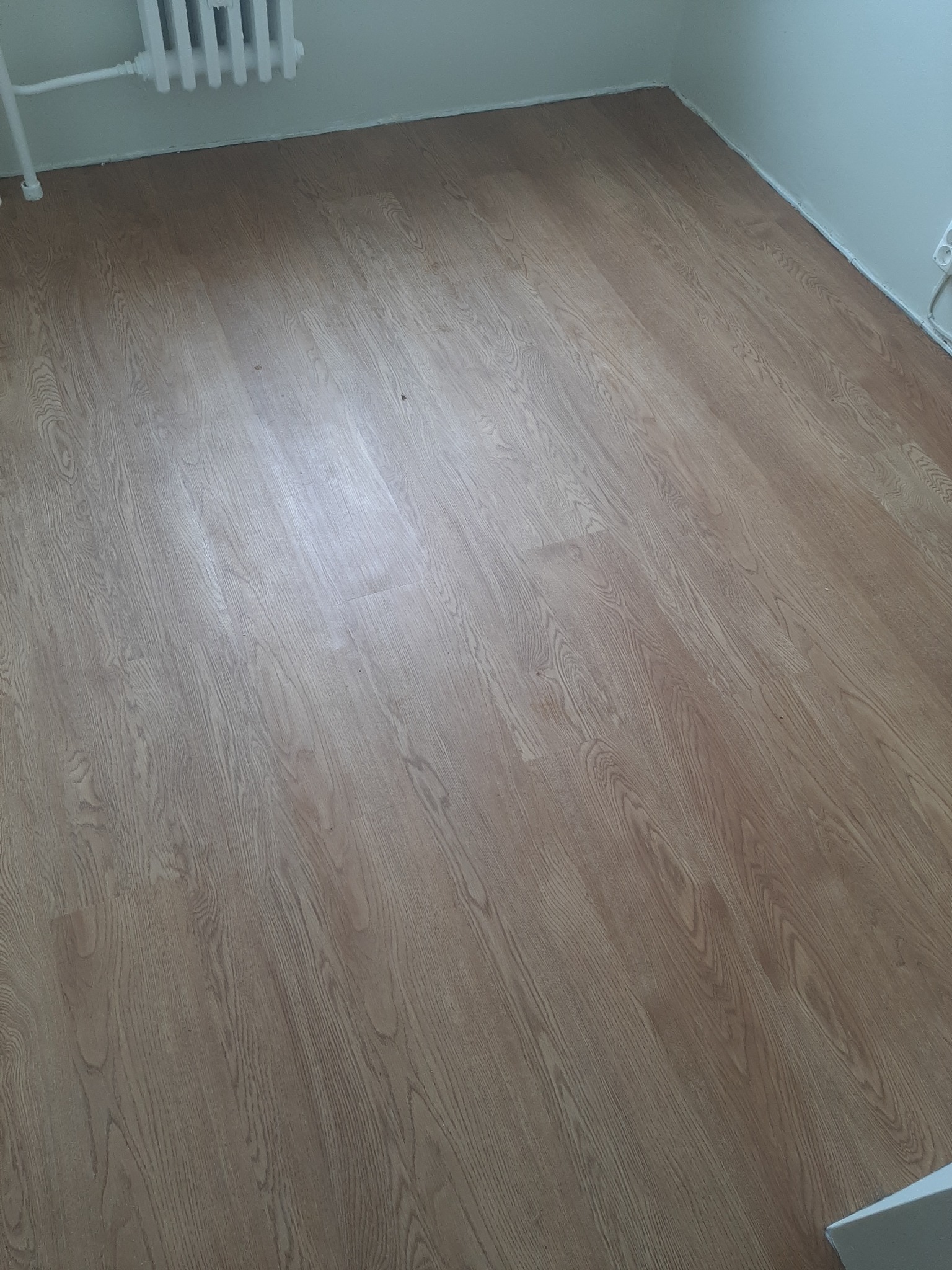 Nowo położona podłoga panelowa o wzorze jasnego drewna, widoczny fragment grzejnika i biała listwa przypodłogowa.