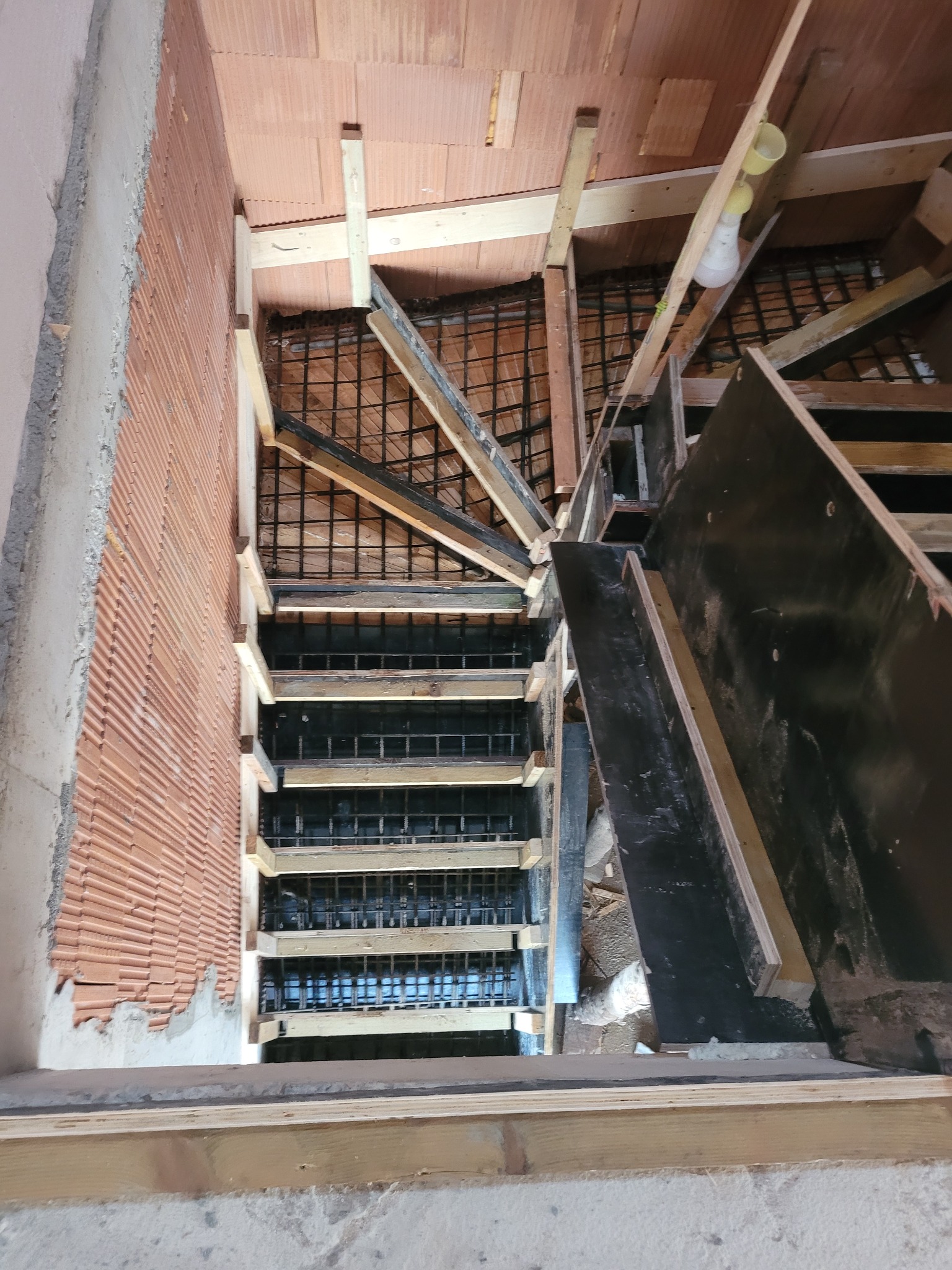 Wnętrze w trakcie budowy schodów z betonowymi stopniami, widoczne zbrojenie i szalunek, surowa cegła na ścianie, wisząca żarówka.