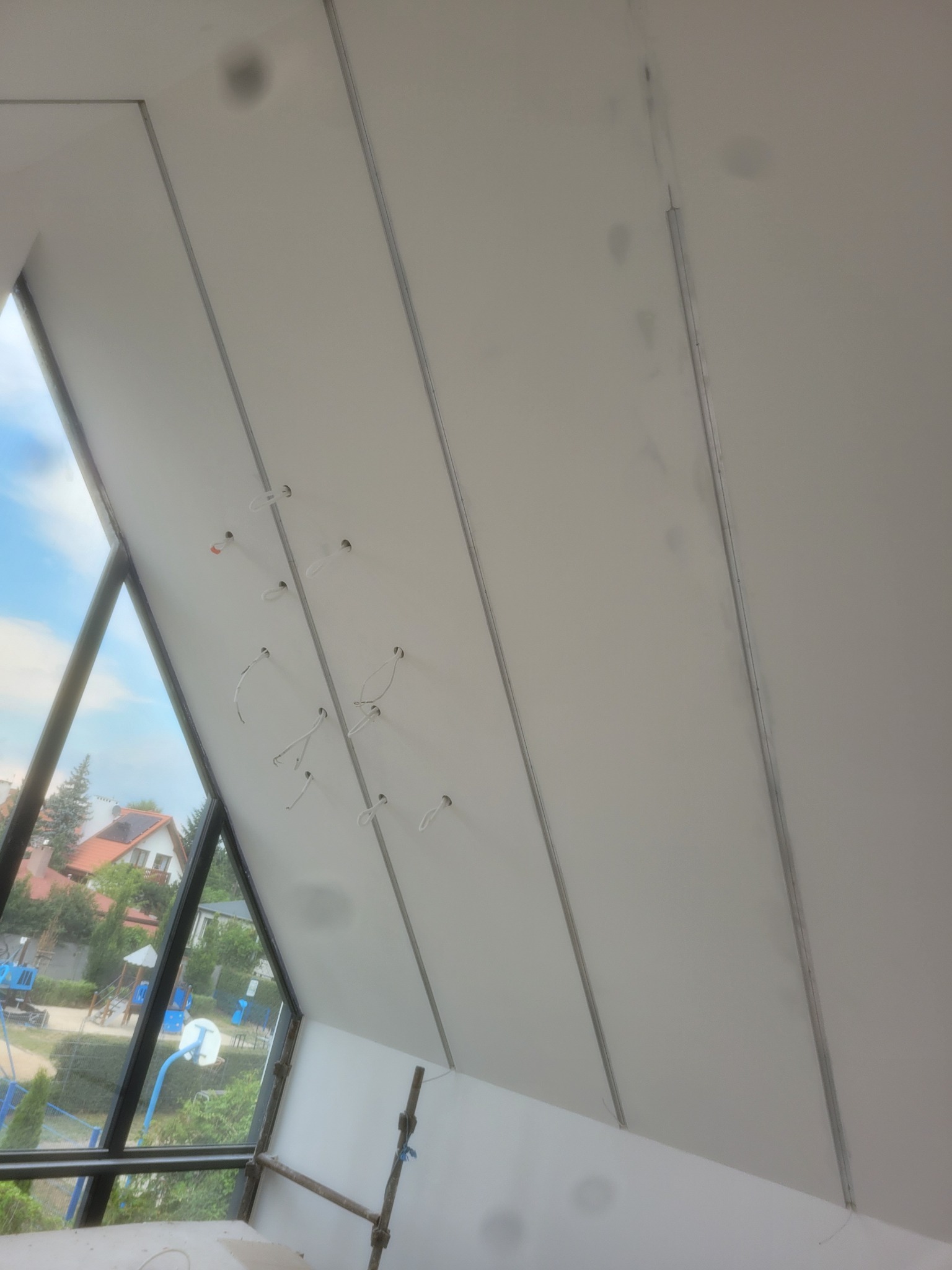 Skośny sufit poddasza w trakcie montażu płyt kartonowo-gipsowych, widoczne profile, otwory na oświetlenie i fragment widoku na zewnątrz przez okno.